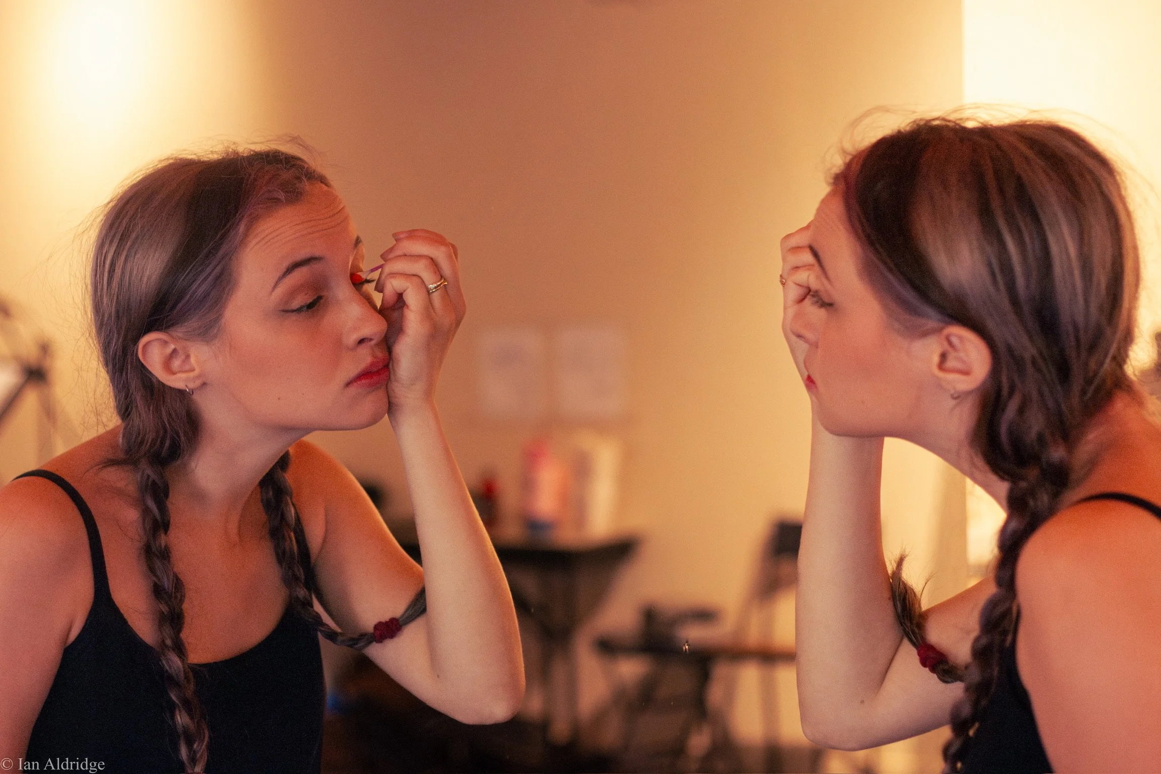 Two women with braided hair sitting and looking at each other, one applying makeup to the other's eye in a warmly lit room.