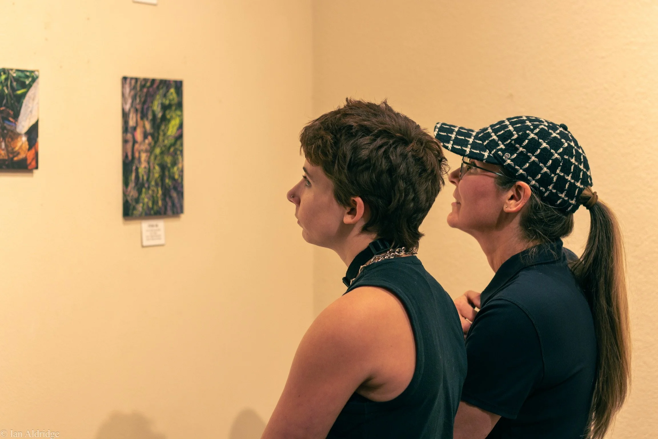 Two women, one with short curly hair and a nose piercing and the other with long hair tied back, are looking at paintings on a beige wall in an art gallery. One woman wears glasses and a patterned cap, and they are both wearing dark sleeveless tops.