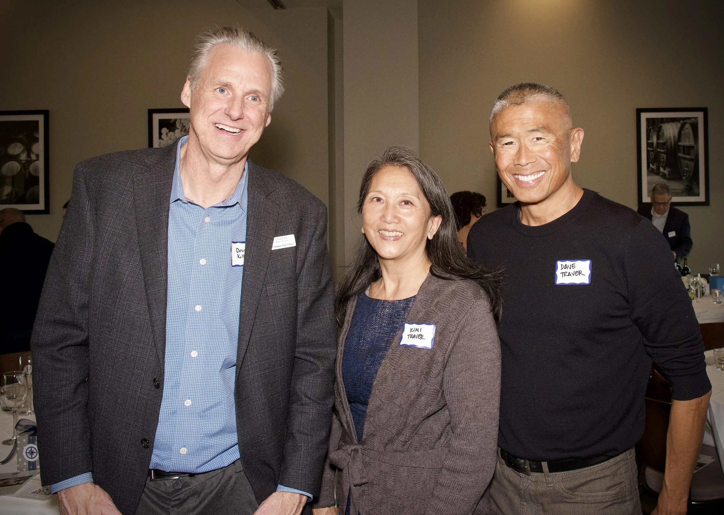 Three smiling adults at a social gathering, two men and one woman, standing close together in an indoor setting with framed black-and-white photographs on the wall behind them.