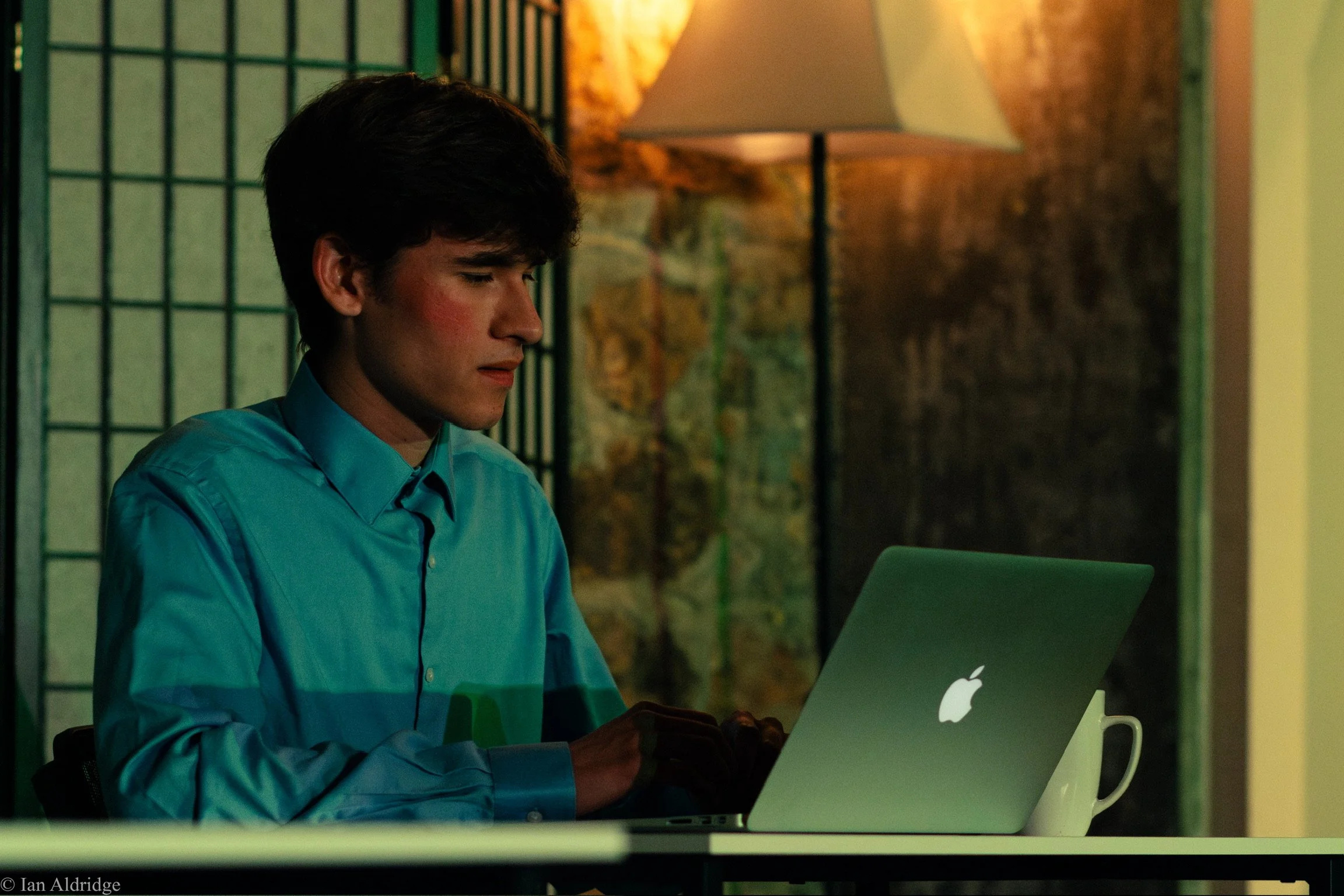 A young man with dark hair wearing a blue shirt working on a laptop in an office setting, with a colorful map and a standing lamp in the background.