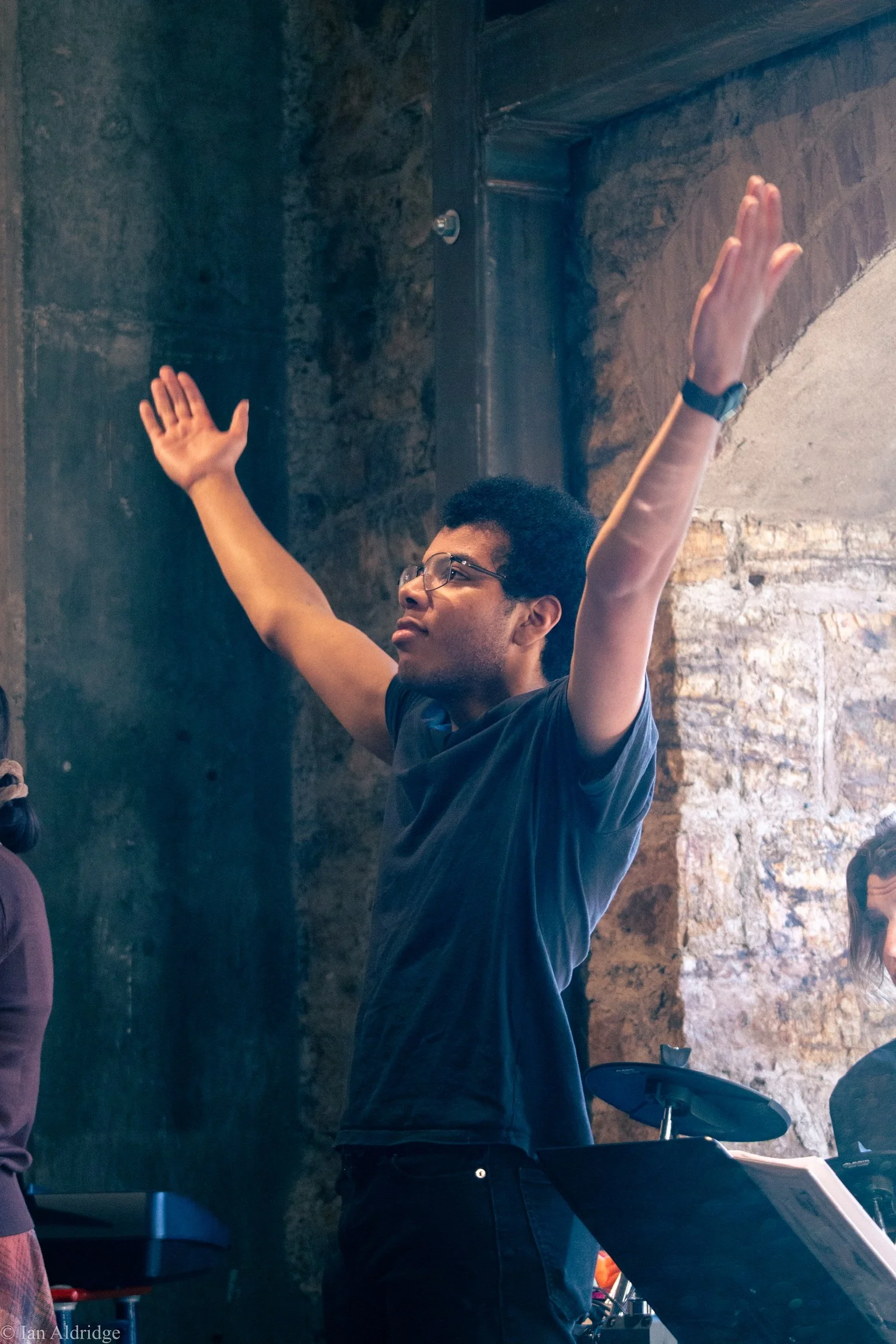 A young man with glasses, wearing a dark t-shirt and a watch, raising his arms while standing next to a digital drum set in a brick-walled room.