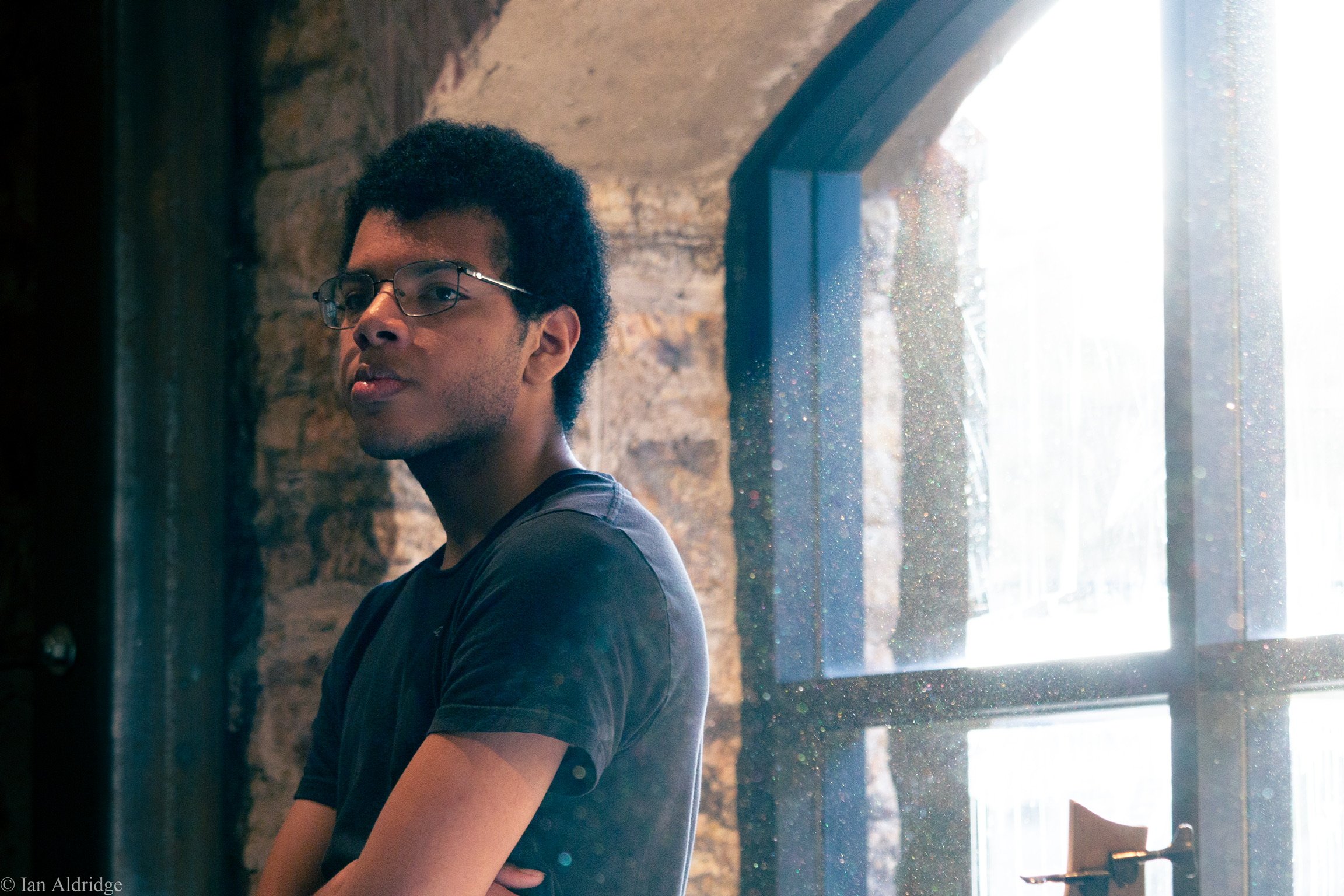 A young man with glasses and curly black hair, wearing a black T-shirt, standing indoors near a large window with bright light. The background has a rustic brick wall.