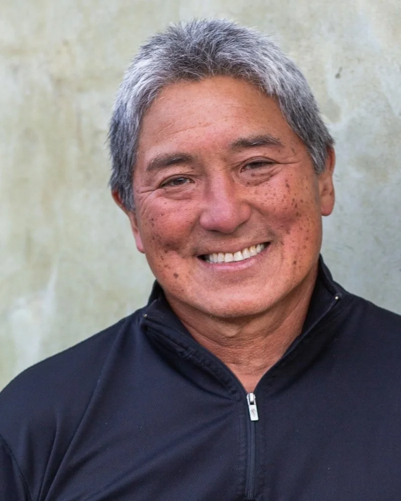 Smiling middle-aged man with gray hair wearing a black jacket, standing outdoors against a plain wall.