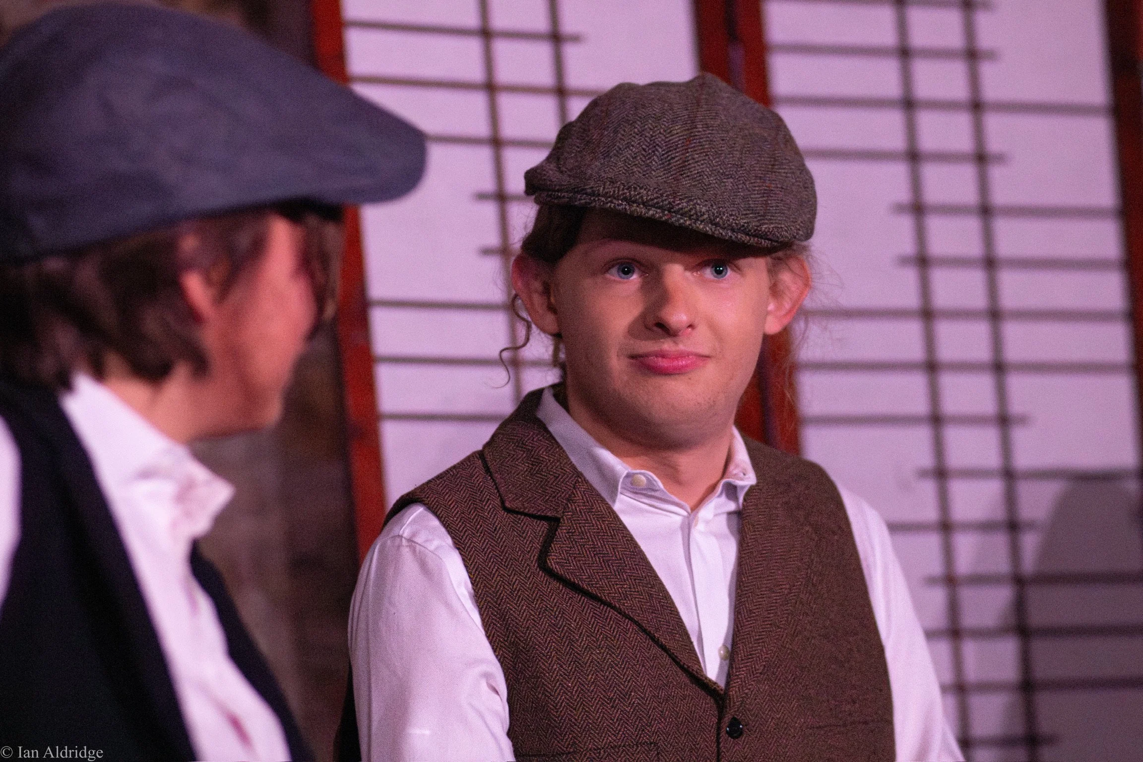 A young man with curly hair wearing a flat cap, white shirt, and brown vest, sitting in front of a shoji screen, looking at a woman with glasses and a bow tie, who is partly visible.
