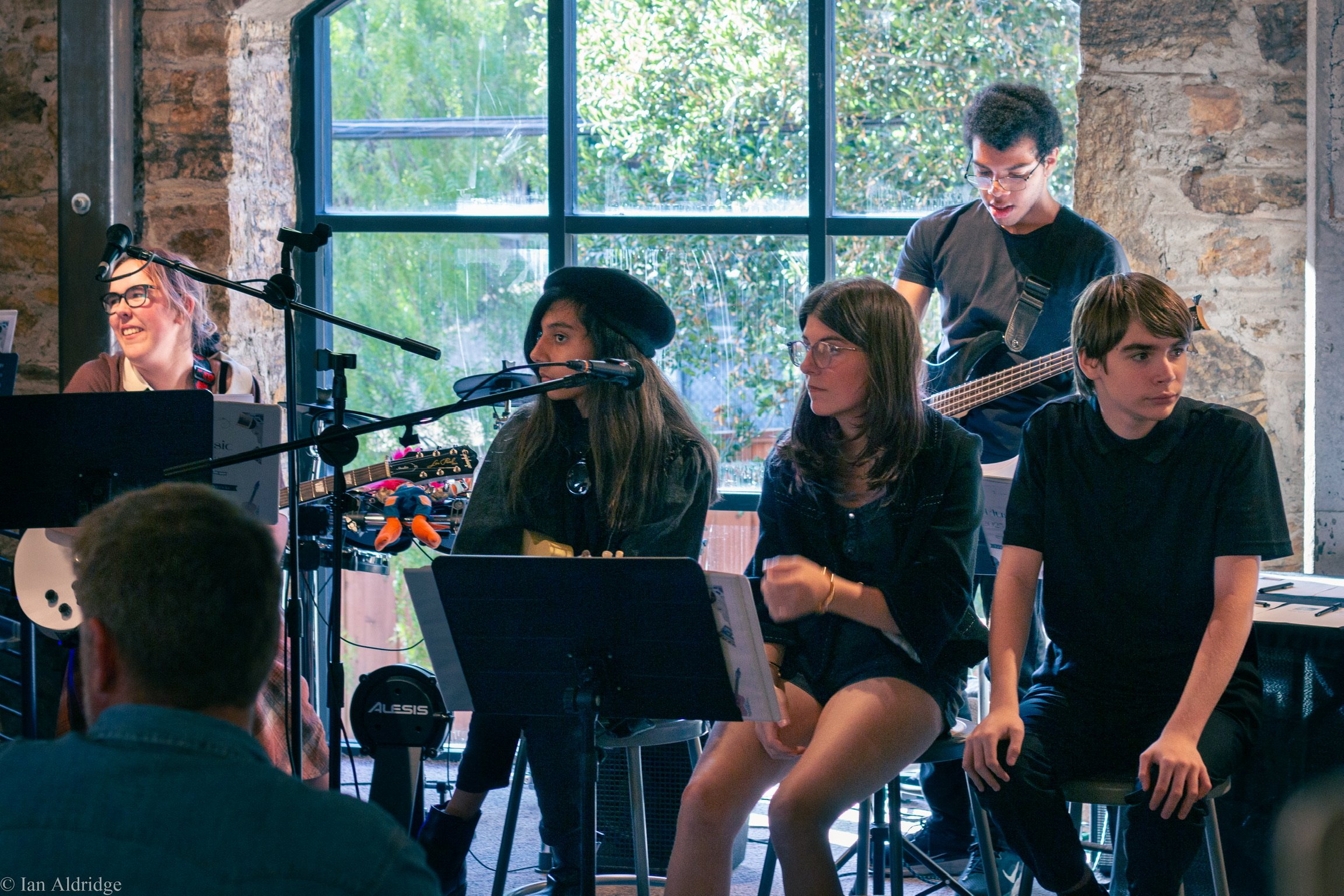 Group of young musicians rehearsing indoors near a large window, with some sitting and others standing, playing instruments and preparing.