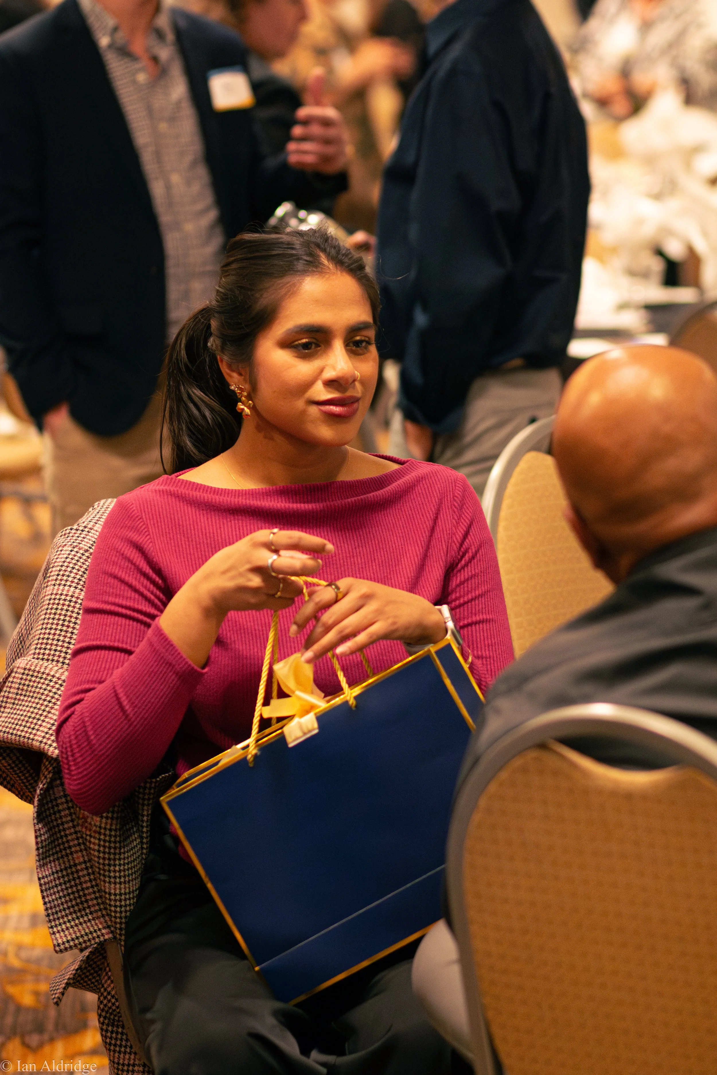 A young woman wearing a pink sweater with dark hair and earrings is holding a blue gift bag with yellow ribbons, sitting and engaged in conversation at a social event. People are standing and talking in the background.