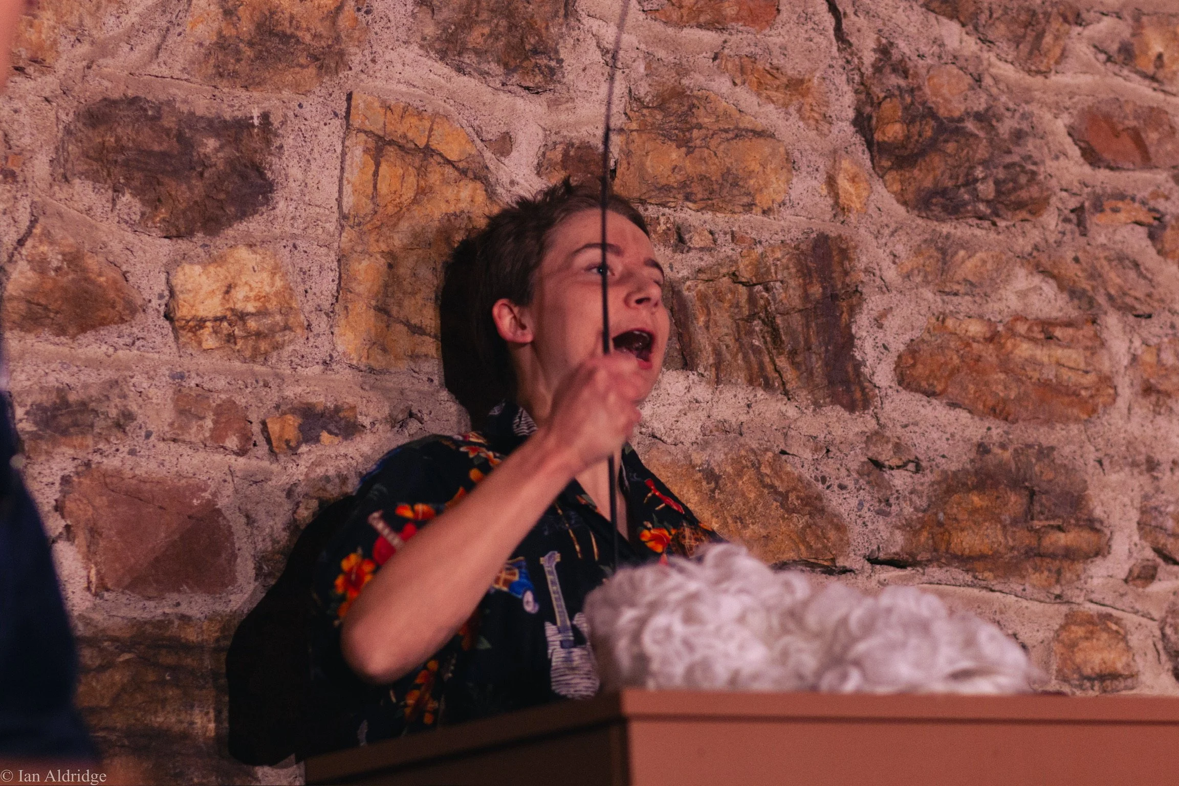 A person with short dark hair and a floral shirt is sitting against a stone wall, holding a black rope or string near their face, with a surprised or playful expression.