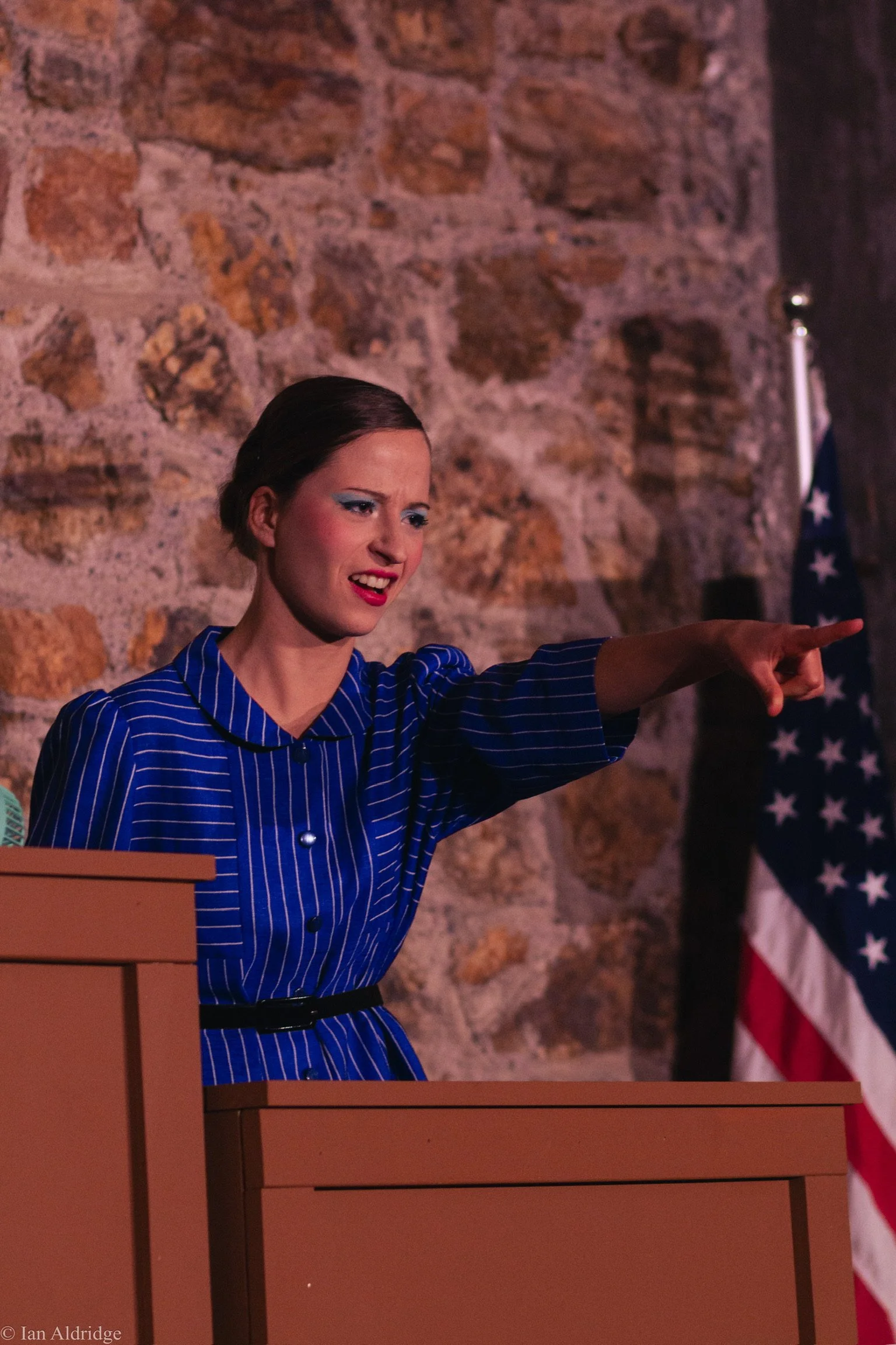 A woman with short dark hair and makeup wearing a blue striped dress is pointing to her right against a background of blocks and an American flag.