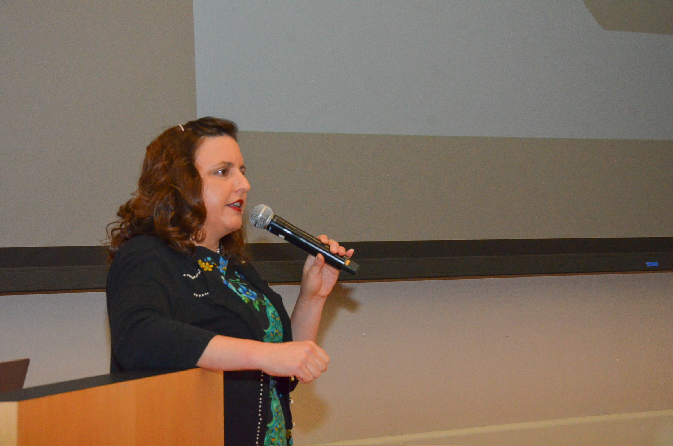 A woman with curly red hair, wearing a black jacket and floral blouse, holding a microphone and speaking in front of a white projection screen.