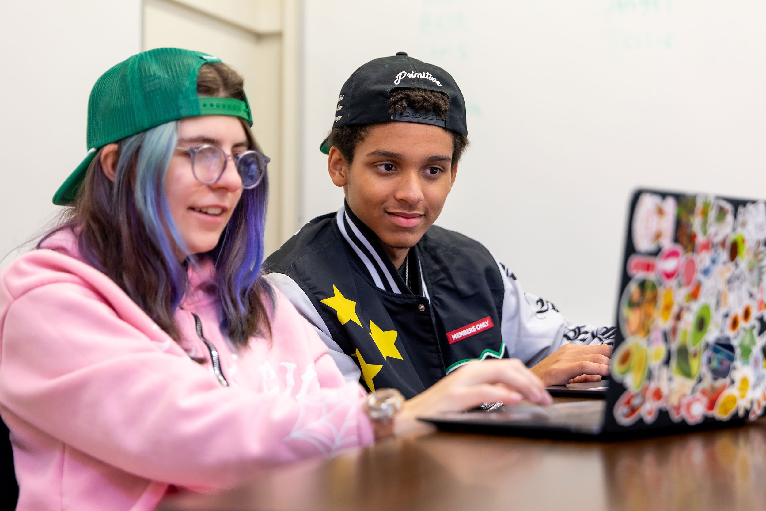 Two students work on their sticker-covered laptops. Both are smiling.