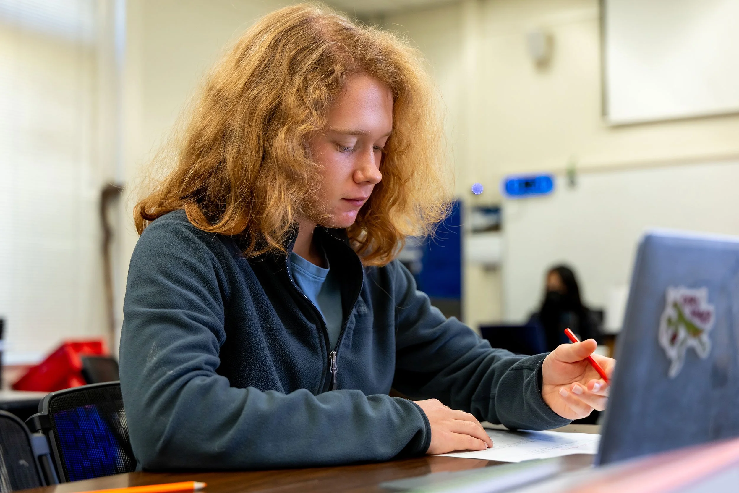 A young person with curly red hair working at a table, writing on paper with a red pen, with a laptop open in front of them in a classroom or study area.