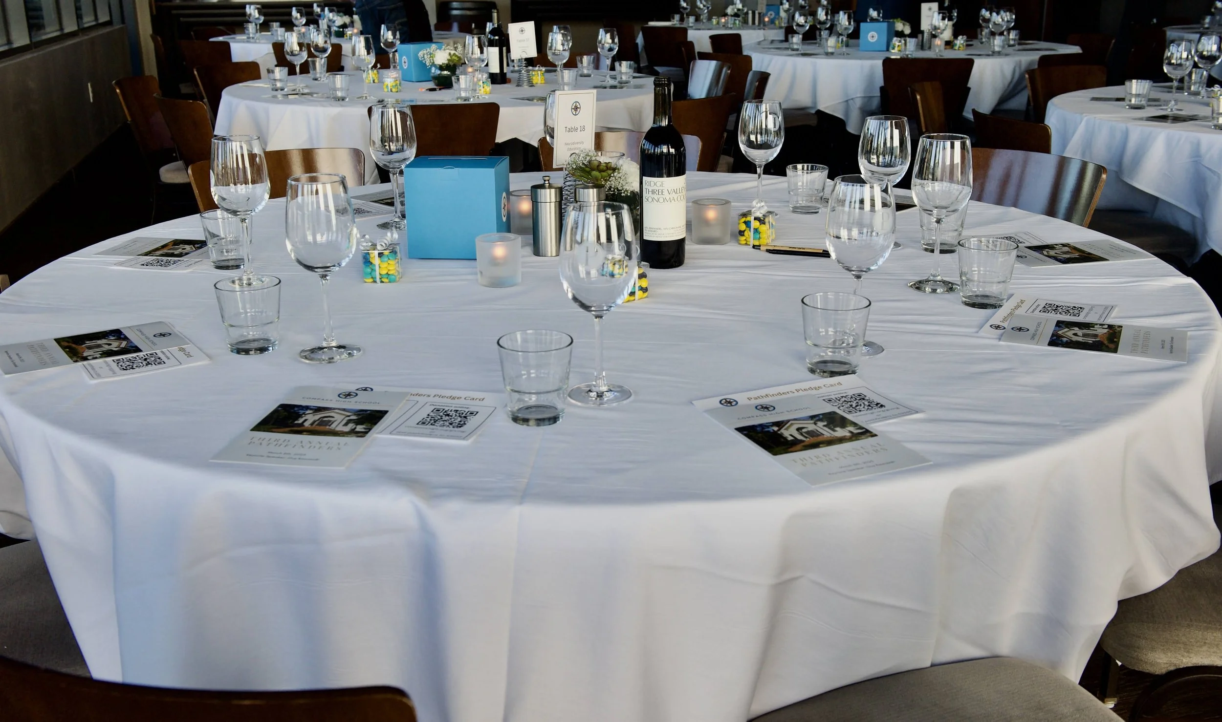 A round banquet table with a white tablecloth set for a formal event, featuring multiple wine glasses, water glasses, a bottle of wine, candles, and place cards with QR codes, in a well-lit room with other similarly set tables in the background.