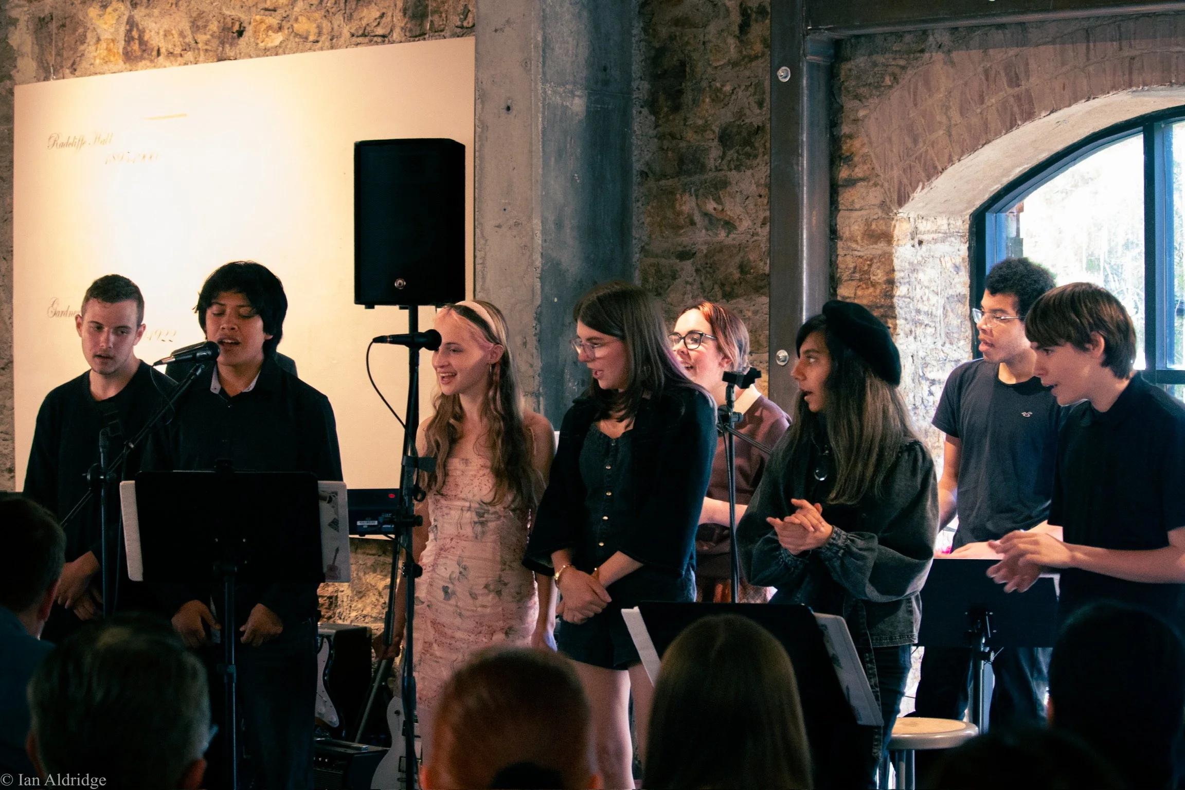 A group of people standing together in a room with stone walls and arched windows, some are singing or speaking into microphones, others appear to be listening or preparing to perform, with a music stand and sound equipment visible.