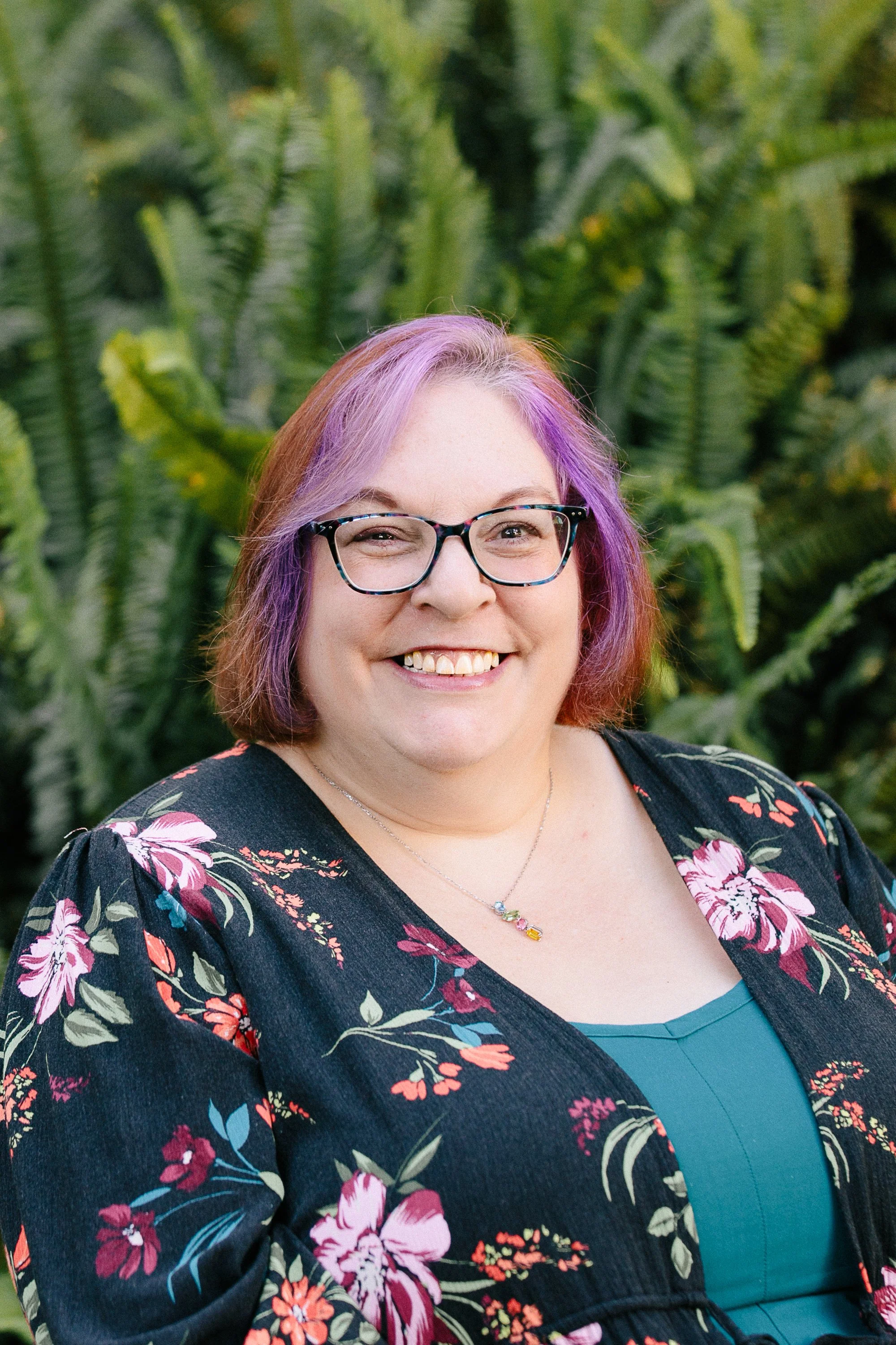 Smiling woman with purple and orange hair, glasses, floral dress, and a necklace, standing outdoors in front of lush green plants.