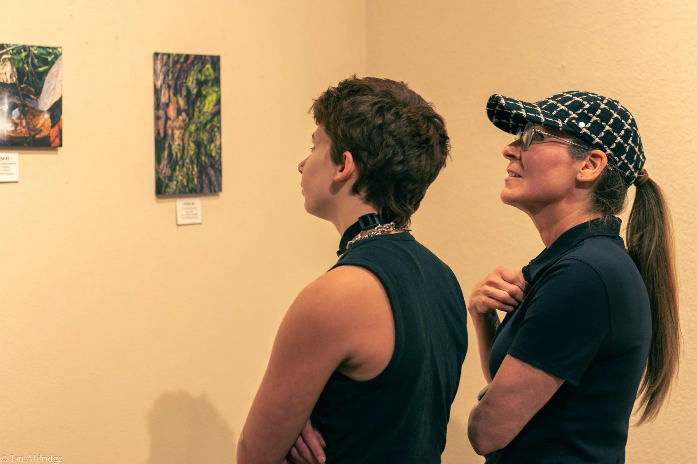 Two women are viewing artwork in a gallery. One woman has short brown hair and is wearing a black sleeveless top with a choker. The other woman has long hair tied back, is wearing glasses, a patterned cap, and a dark top. They are standing side by si