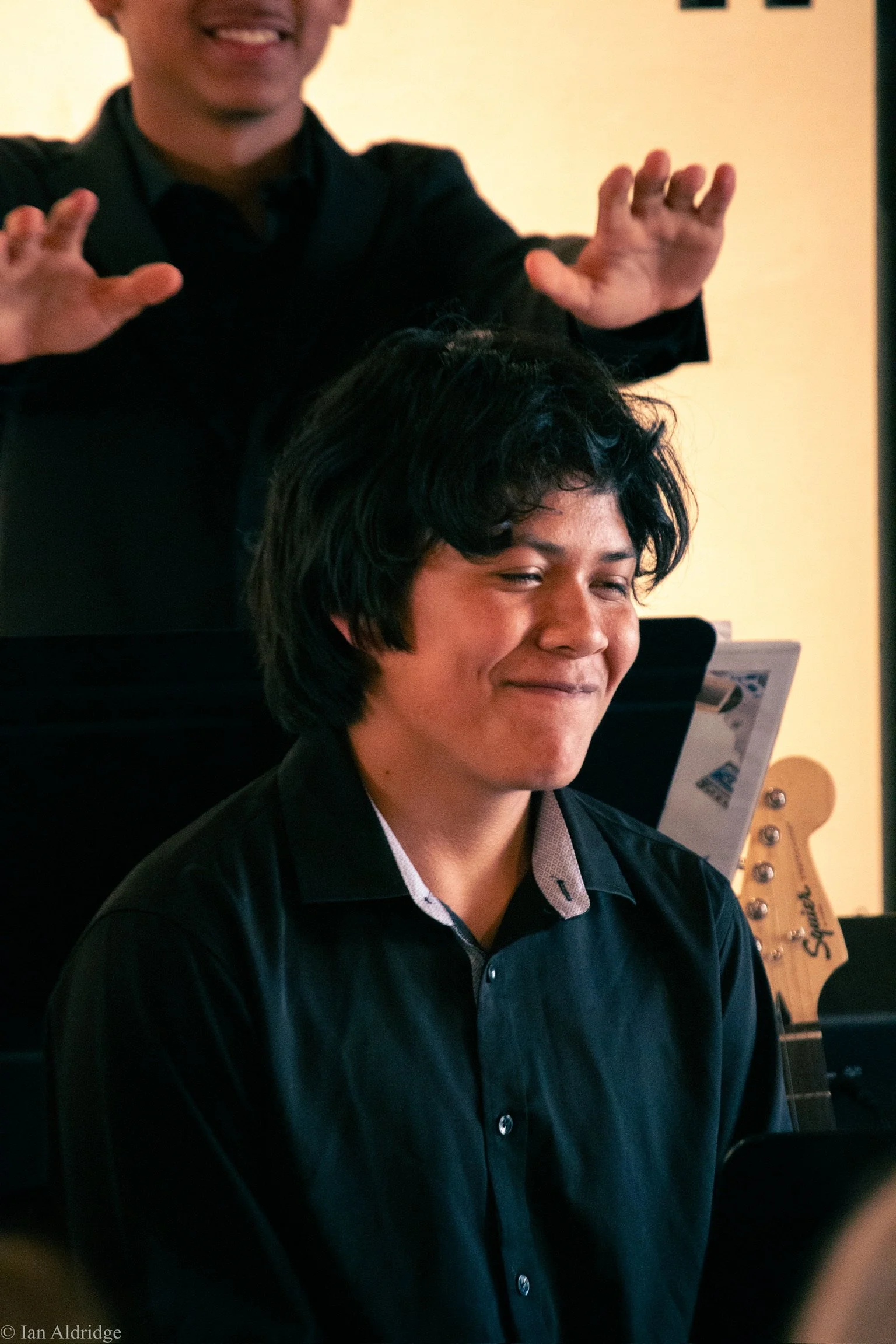 A person wearing a black jacket and shirt, smiling with their eyes closed, with a guitar visible in the background.