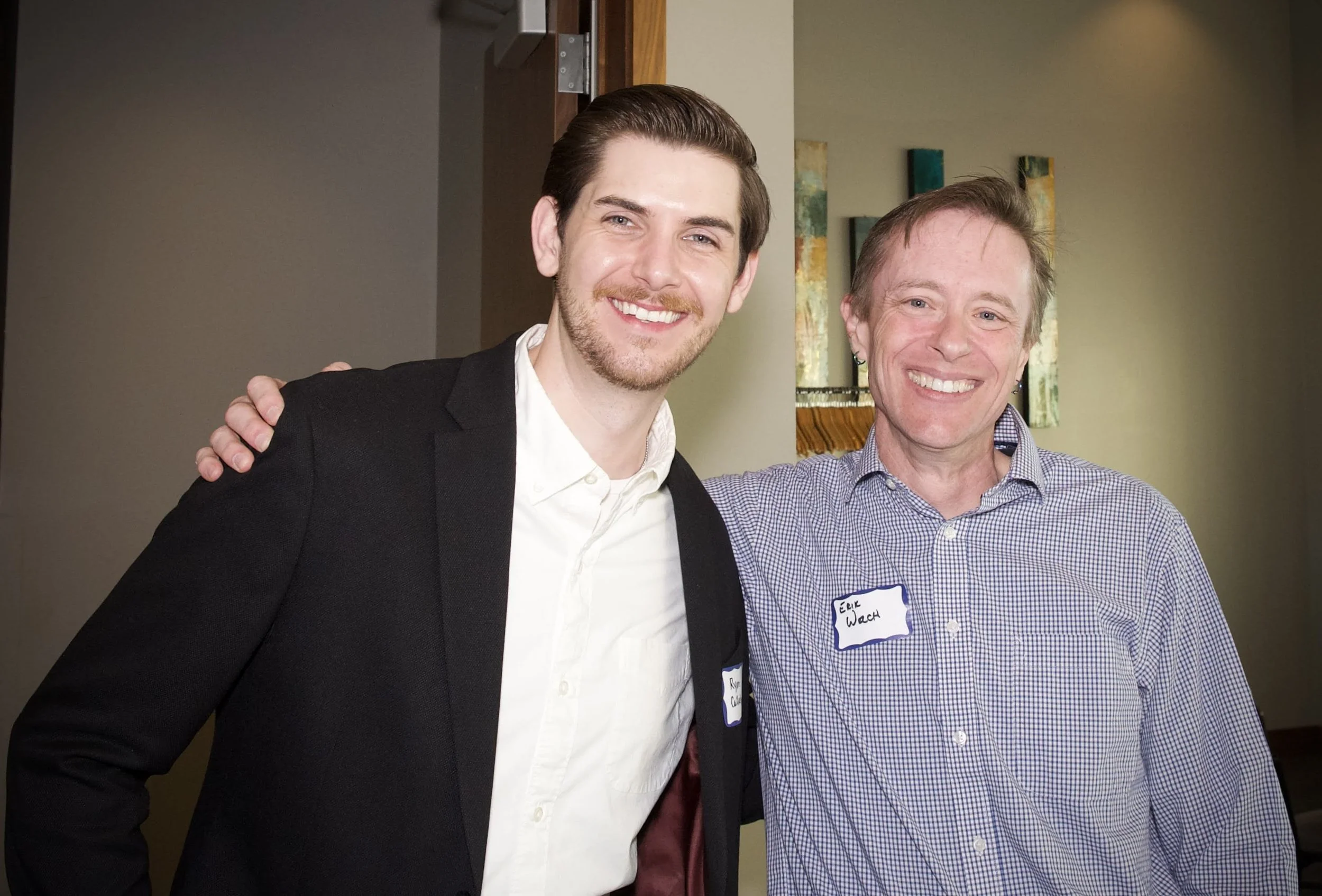 Two men posing together and smiling in an indoor setting. The man on the left has dark hair, a beard, and is wearing a black blazer and white shirt. The man on the right has light brown hair, earrings, and is wearing a checkered shirt with a name tag
