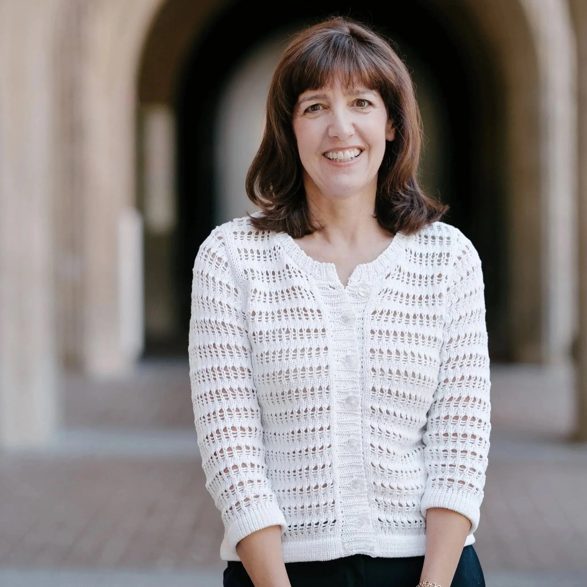 A woman with shoulder-length brown hair and bangs smiling, wearing a white crochet sweater and a gold bracelet, standing outdoors in front of blurred archways.