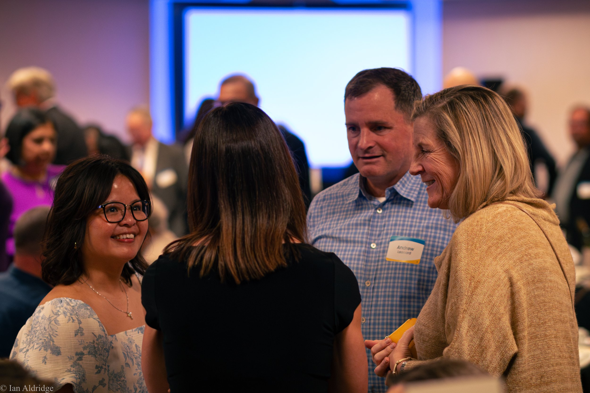 Group of people engaging in conversation at a professional event or conference, with a large screen in the background.