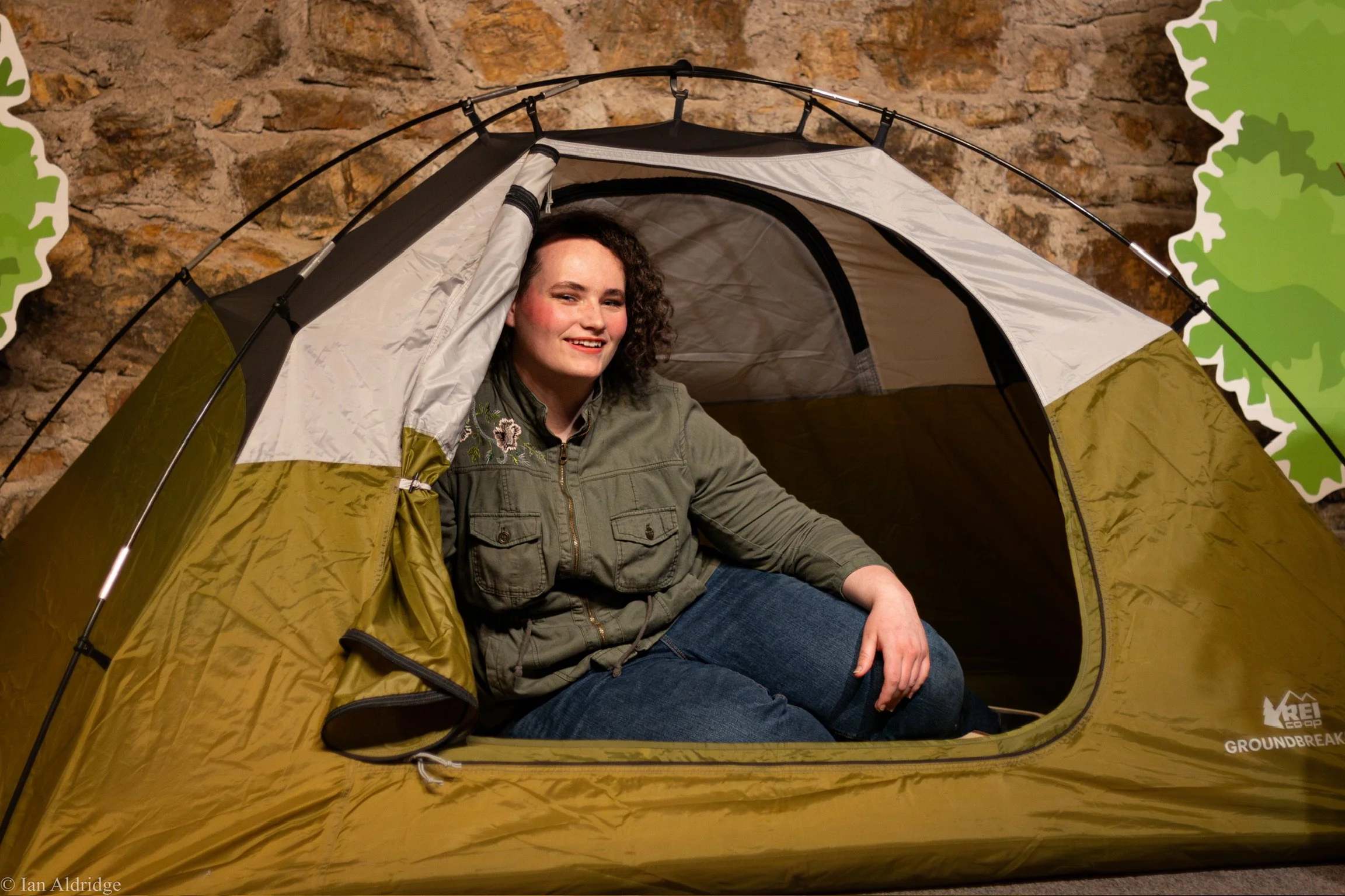 Woman sitting inside a camping tent with a stone wall background, smiling at the camera.