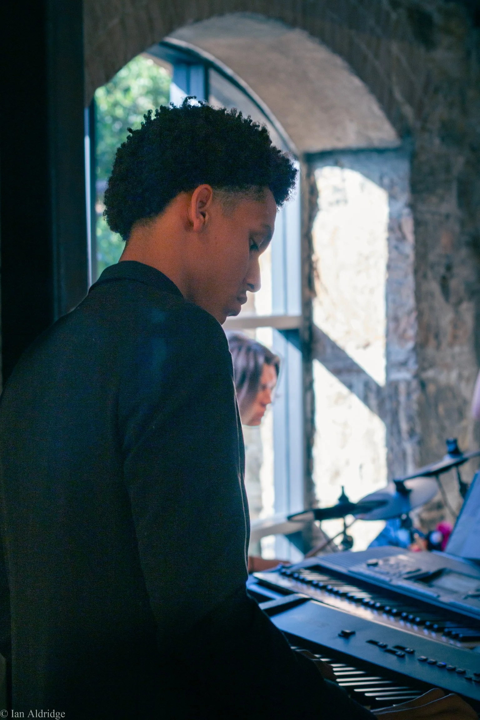 A person with short curly hair playing a keyboard near a window with stone walls and trees outside.