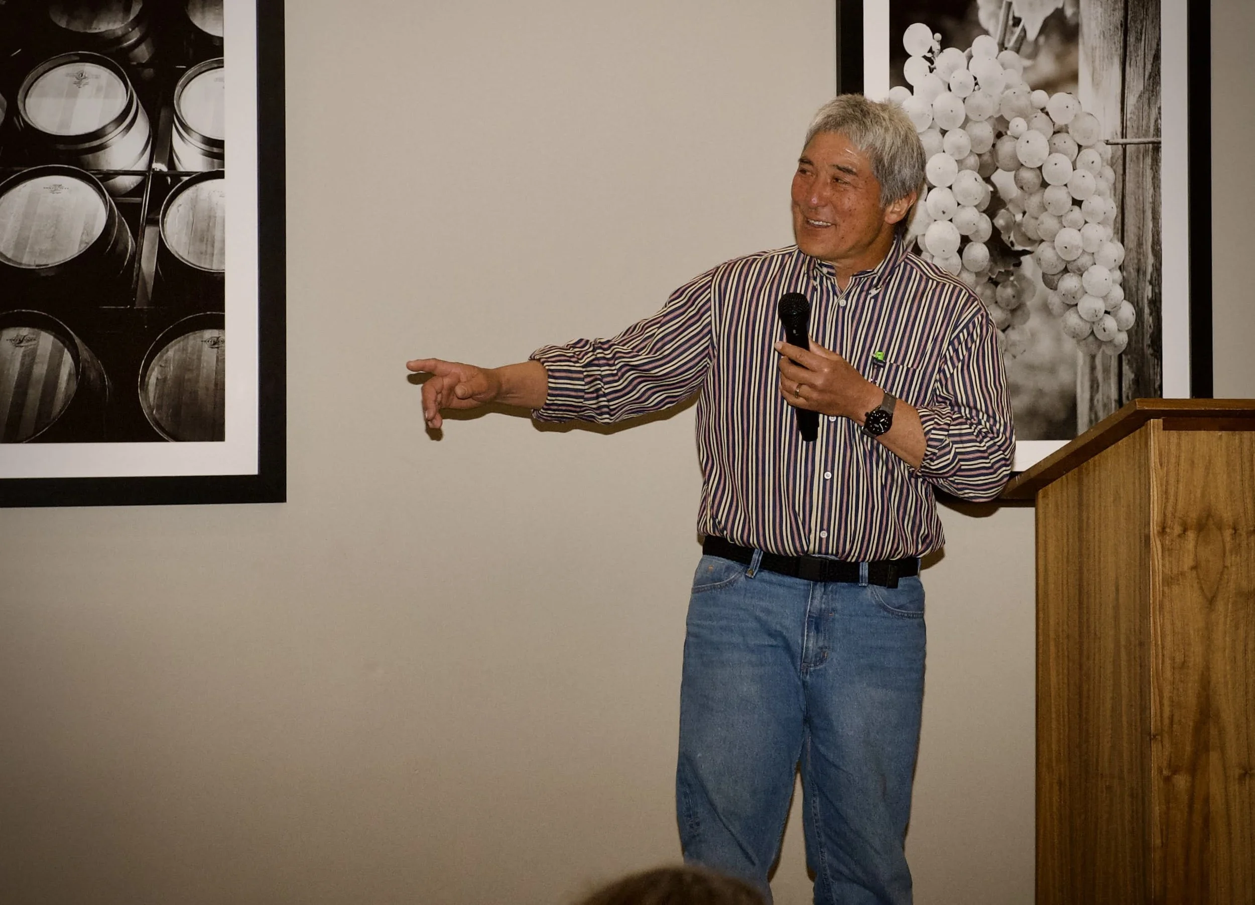 A man with gray hair, wearing a striped shirt and jeans, holding a microphone and giving a presentation, standing next to a podium with a wooden surface, with black and white photographs of grapes and barrels on the wall behind him.