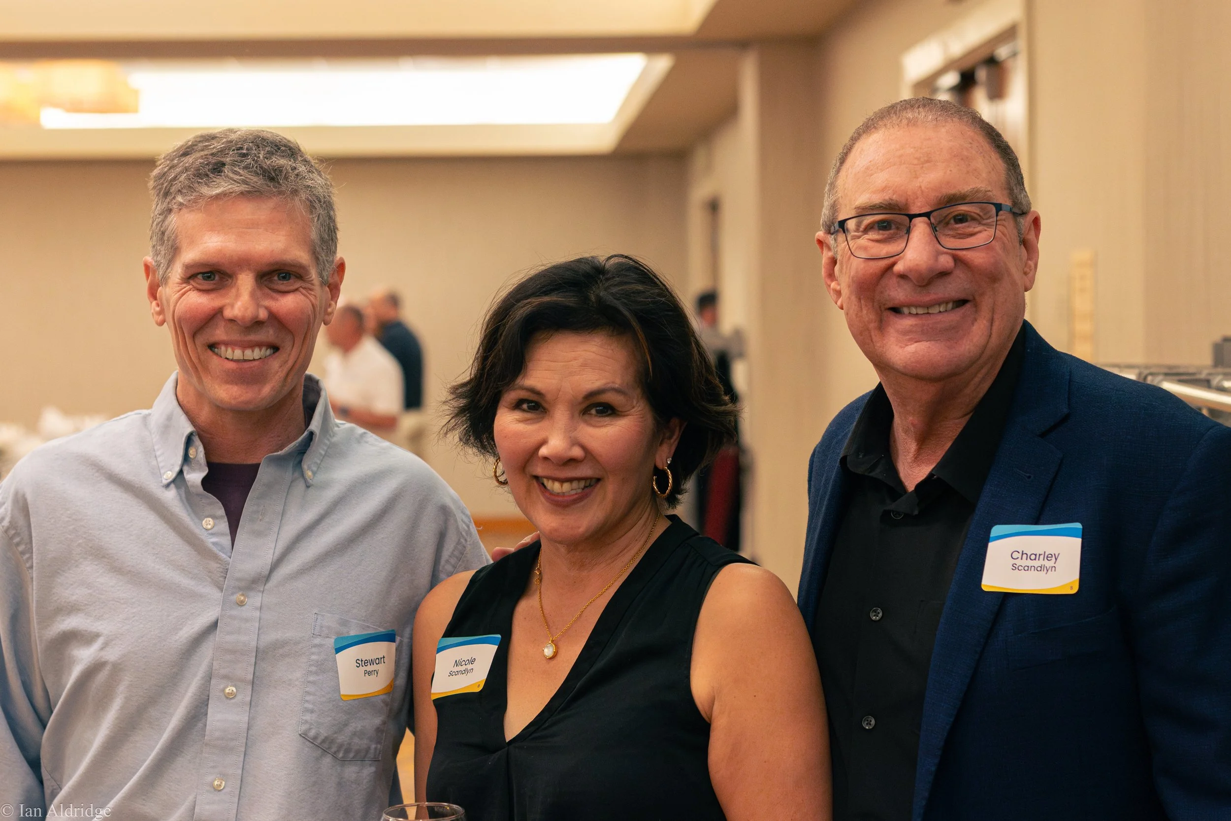 Three people smiling at a social event, two men and one woman, wearing name tags. The background shows a reception area in a hotel or conference center.