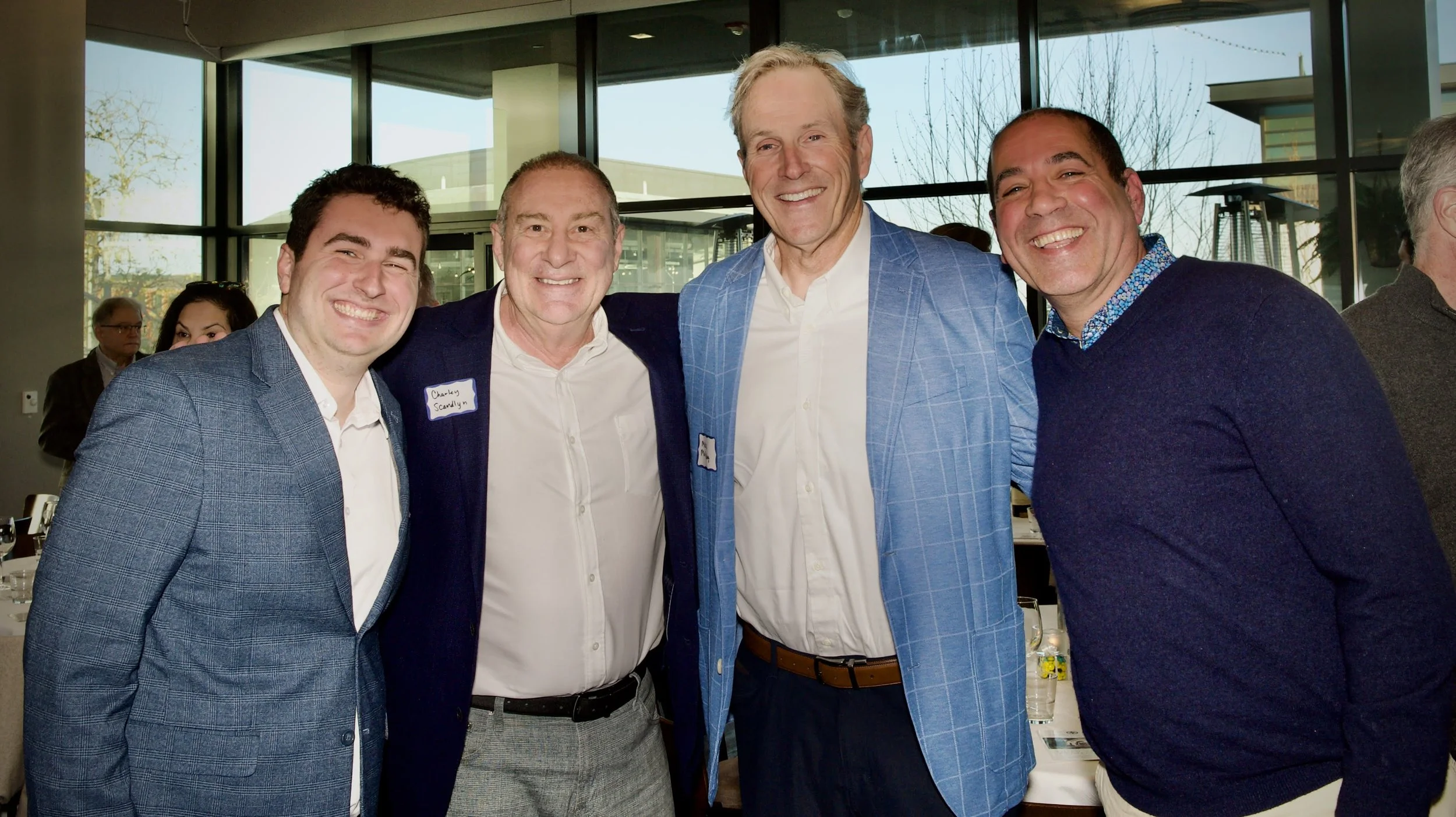 Four men smiling and standing close together at an indoor event, with large windows and a few other people visible in the background.