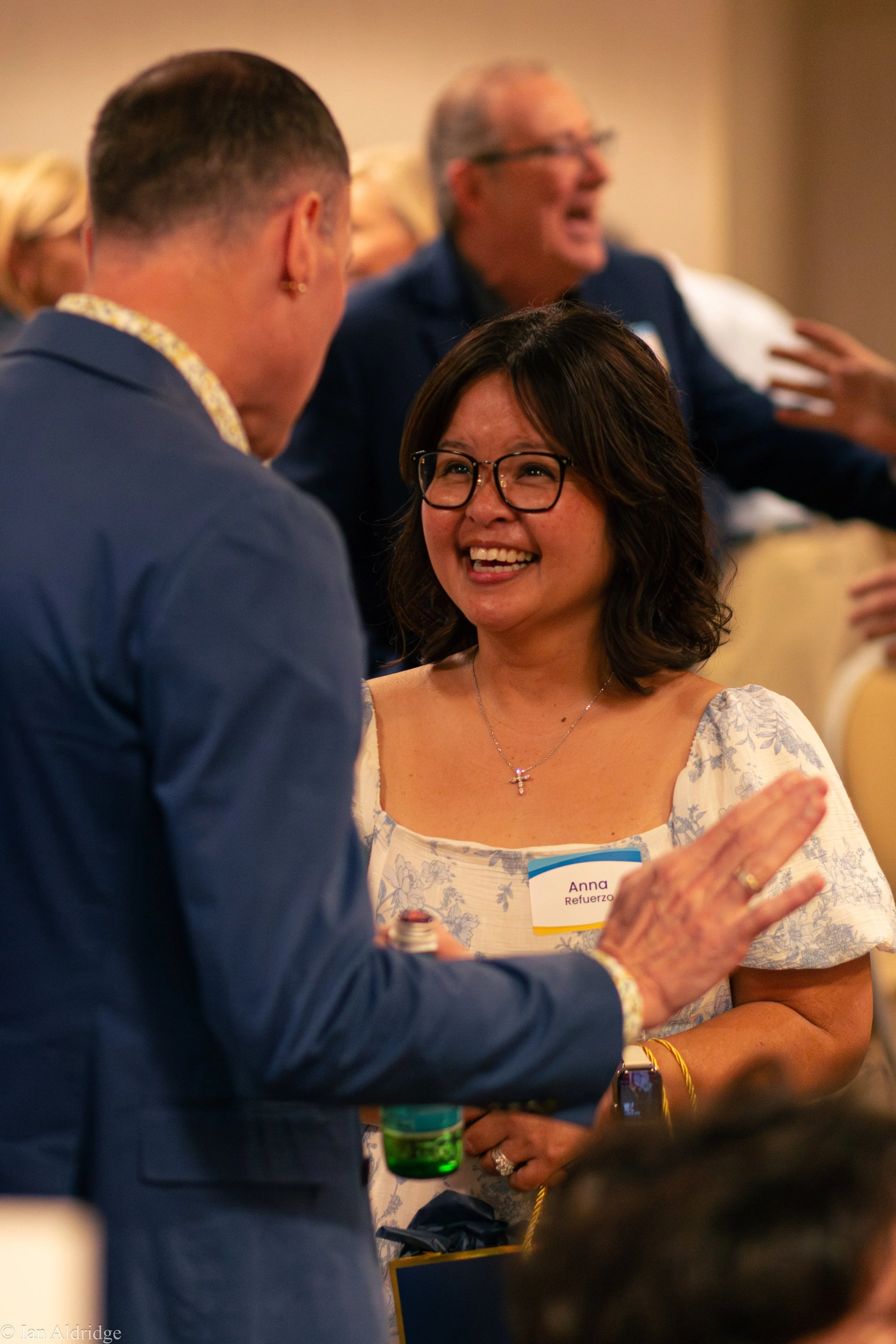 A woman wearing glasses and a white floral dress is smiling and wearing a name tag that reads 'Anna Refuerzo'. She is engaged in conversation with a man in a navy blazer. Other attendees are blurred in the background at what appears to be a social or