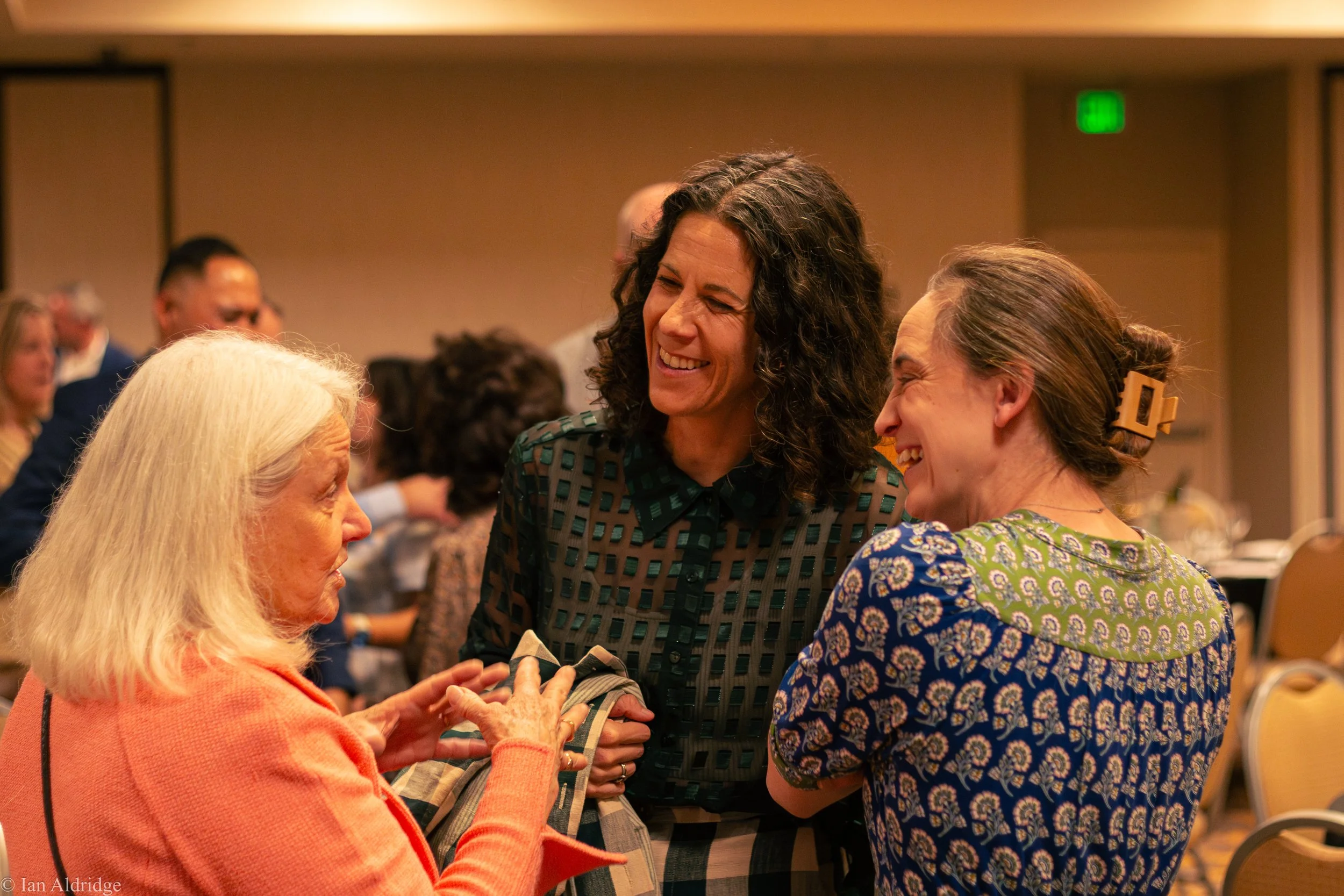 Three women engaged in conversation at a social event, smiling and interacting warmly in a room with other attendees in the background.