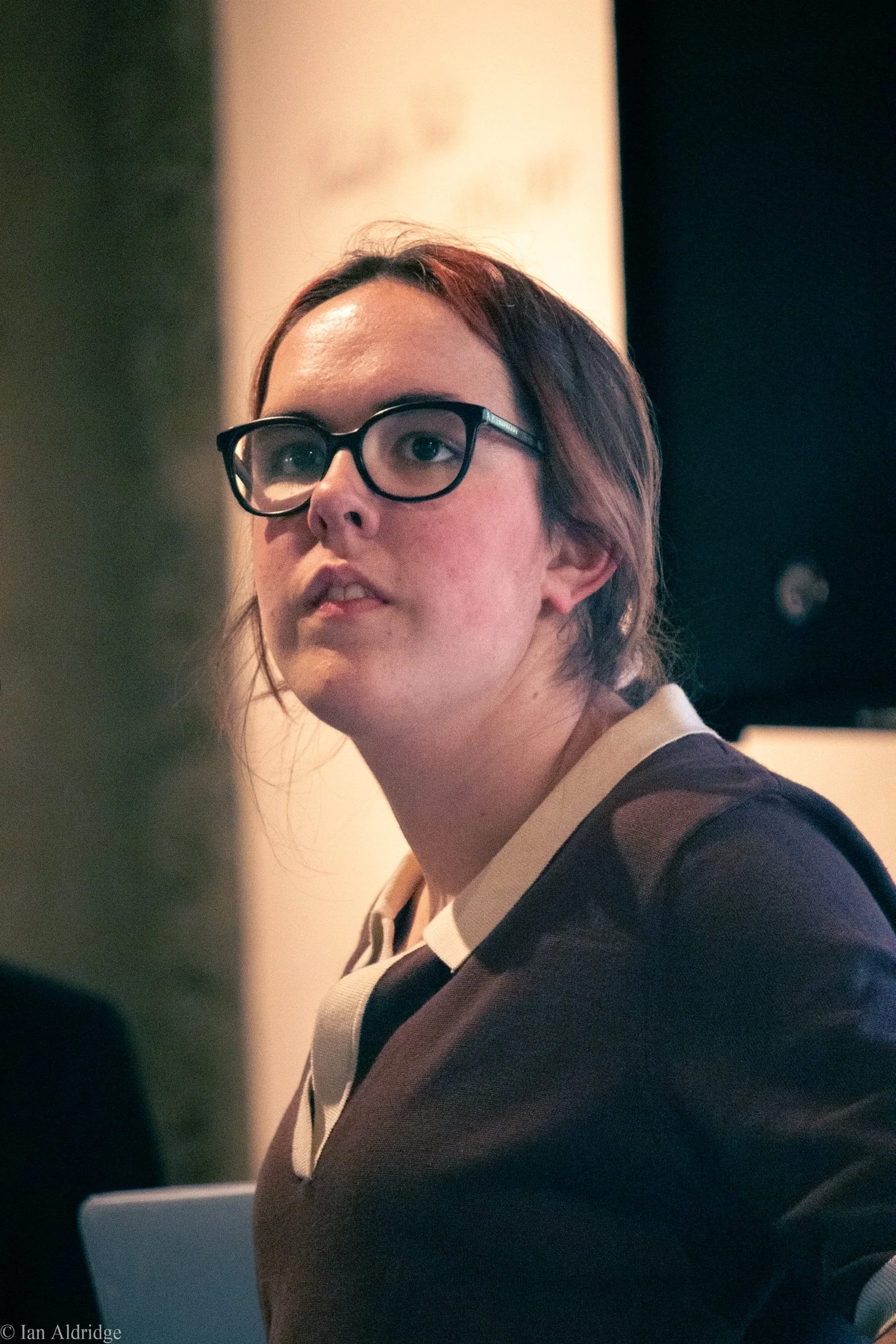 Close-up of a young woman with brown hair and glasses, wearing a dark sweater and light collared shirt, looking to the left.