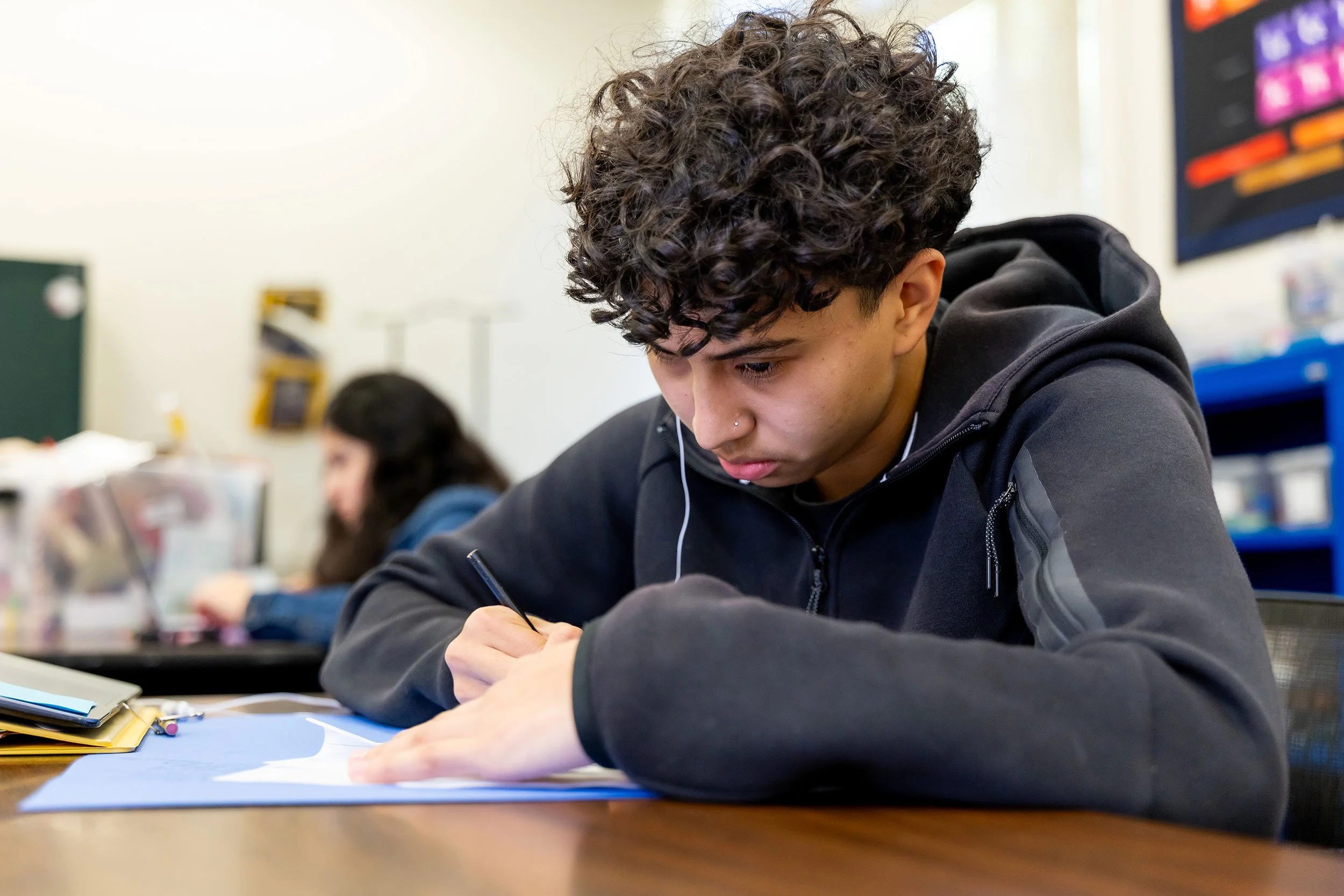 A young man with curly hair and a nose piercing concentrating on writing in a notebook at a desk in a classroom.