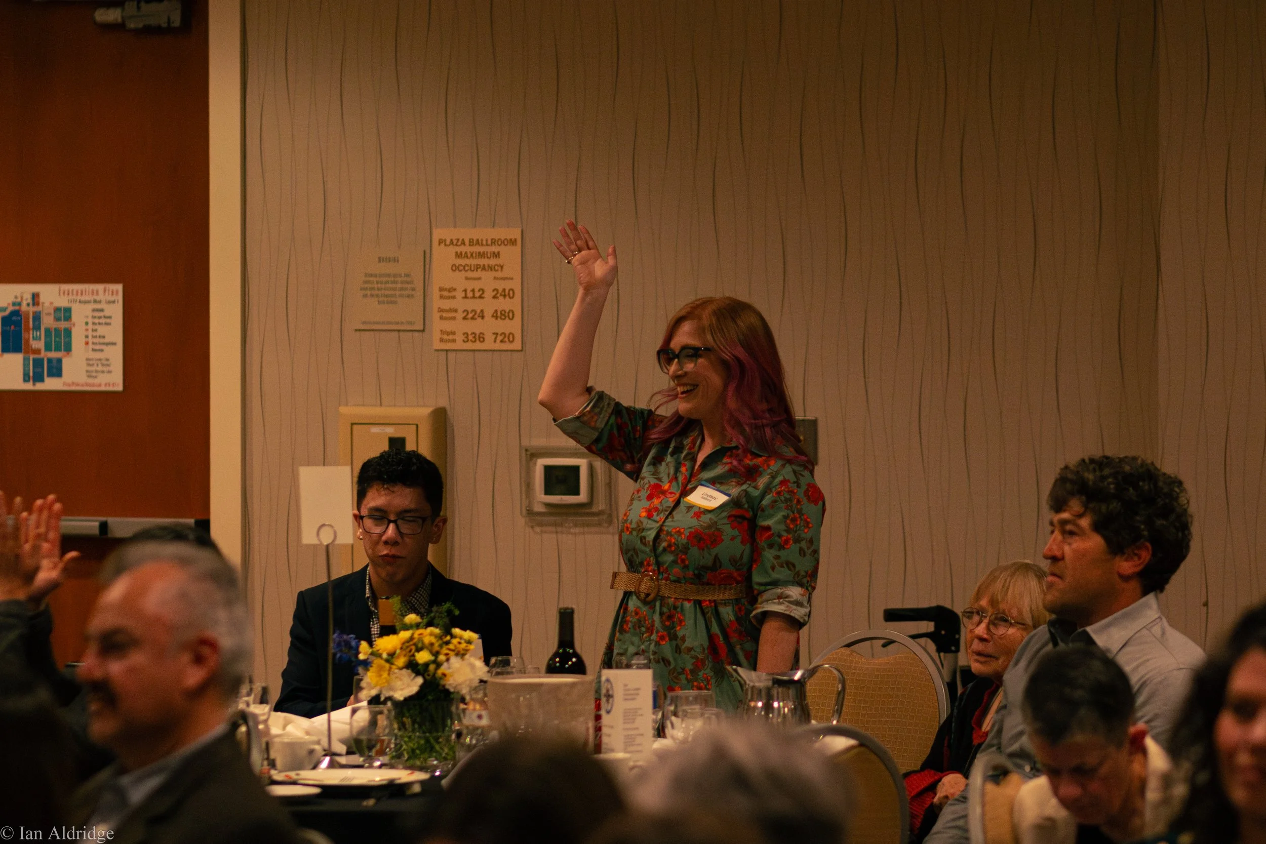 A woman with pink hair and glasses is standing and smiling, raising her hand in a gesture of greeting or participation at a formal event. She is wearing a colorful, floral dress. Several people are seated at tables, and the setting appears to be a ba