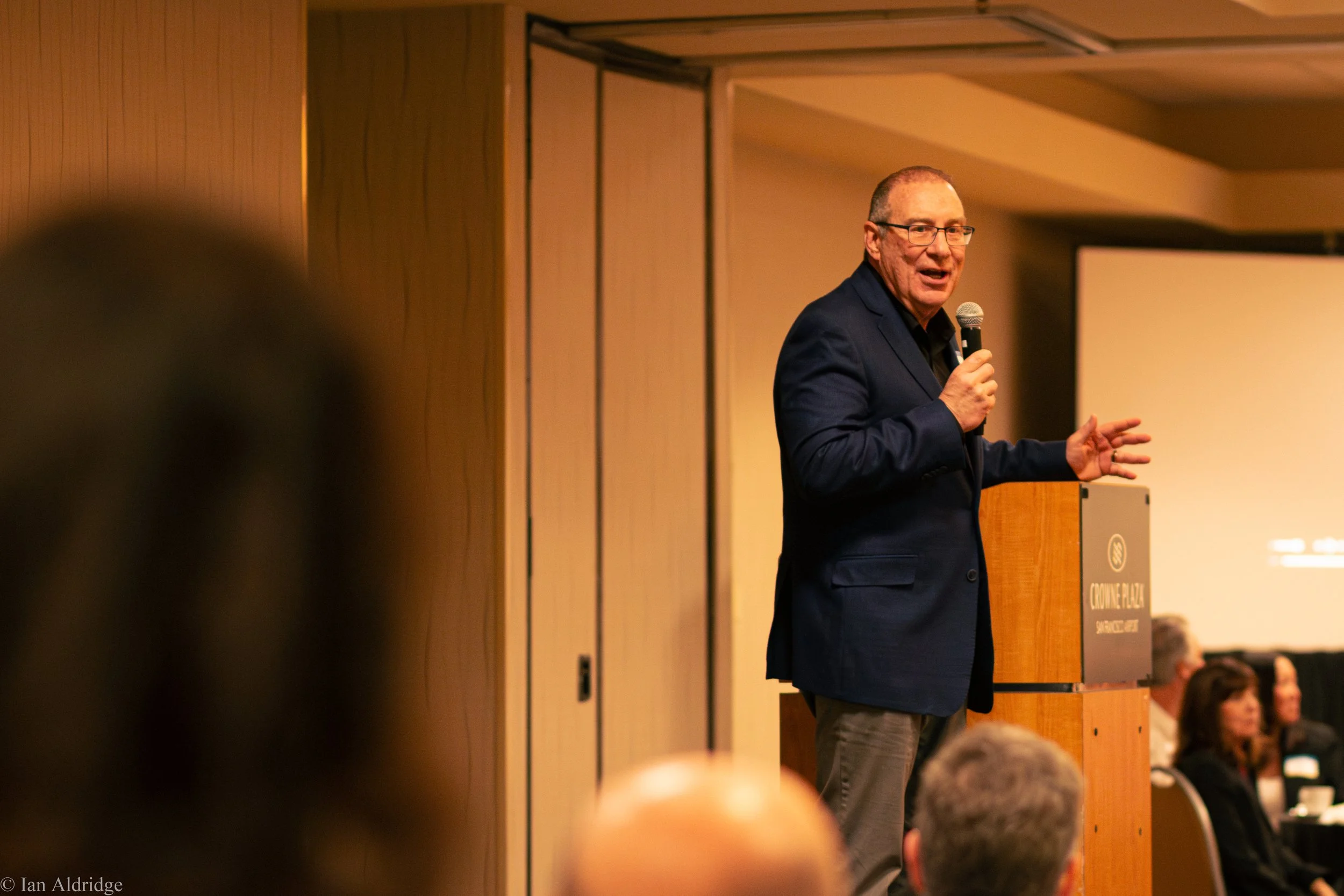 A man in a dark suit holding a microphone and speaking at a conference or seminar, with a group of audience members visible in the foreground and a projection screen nearby.