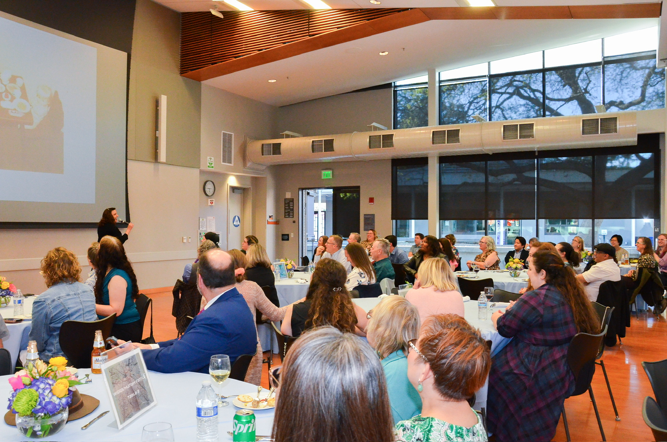 A large group of people sitting at round tables in a banquet hall or conference room, listening to a woman speaking on stage. There are large windows with trees outside, and floral centerpieces on the tables.