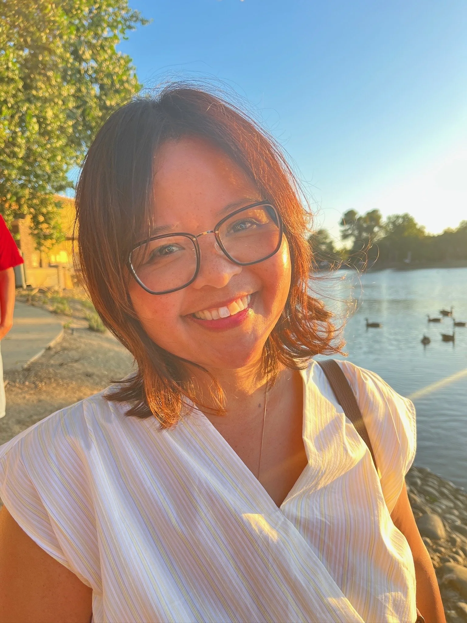 A woman with glasses smiling at the camera during sunset by a body of water with ducks.