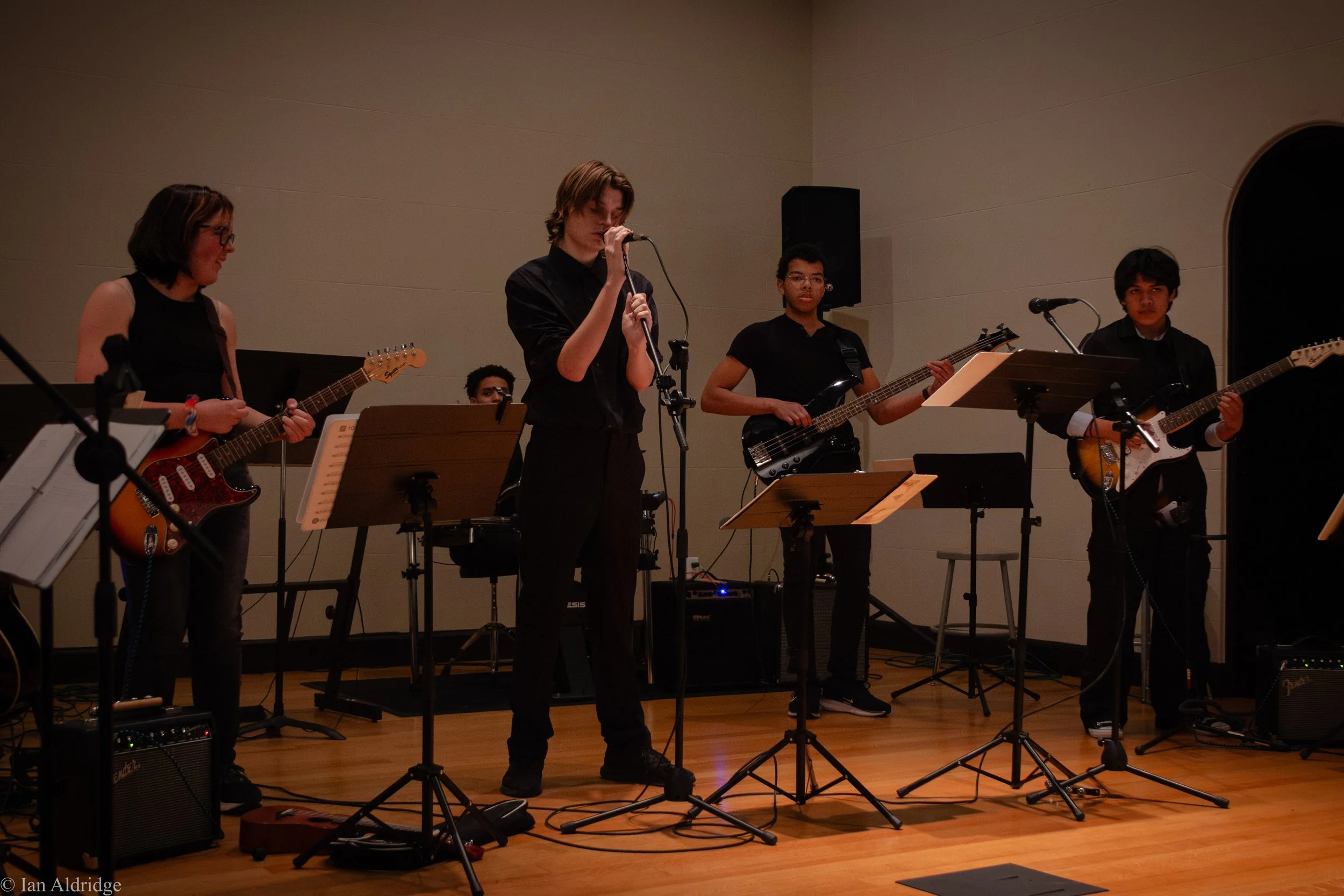 A band of five young musicians performing on stage, with three guitarists, a bassist, and a singer, in a dimly lit room with beige walls.