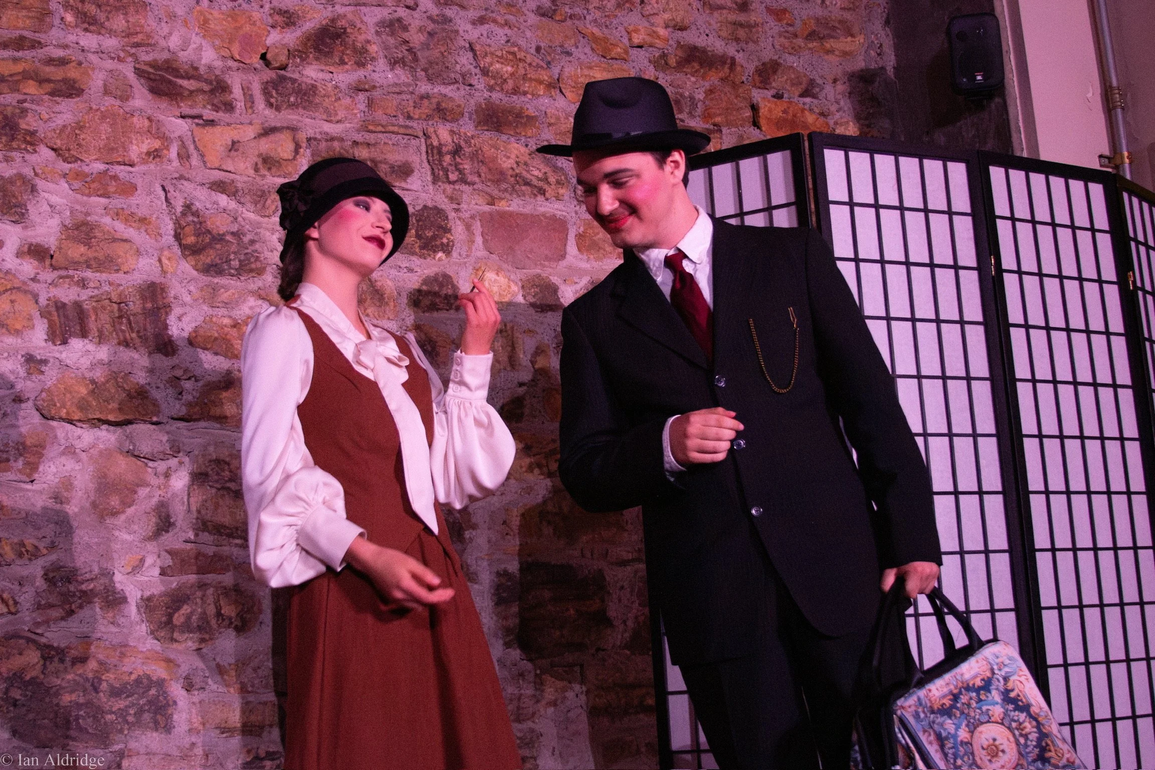 A man in a dark suit and hat and a woman in vintage clothing, with a hat, talking and smiling in front of a brick wall and a folding screen.