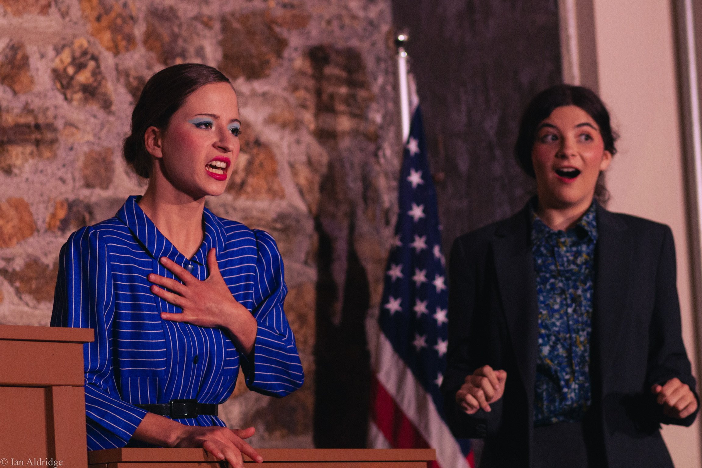 A woman with vintage makeup and a blue pinstripe dress stands with her hand on her chest, speaking passionately. Two other women stand behind her, one with curly hair and a floral shirt, and the other with straight hair and a dark blazer, all in a ro