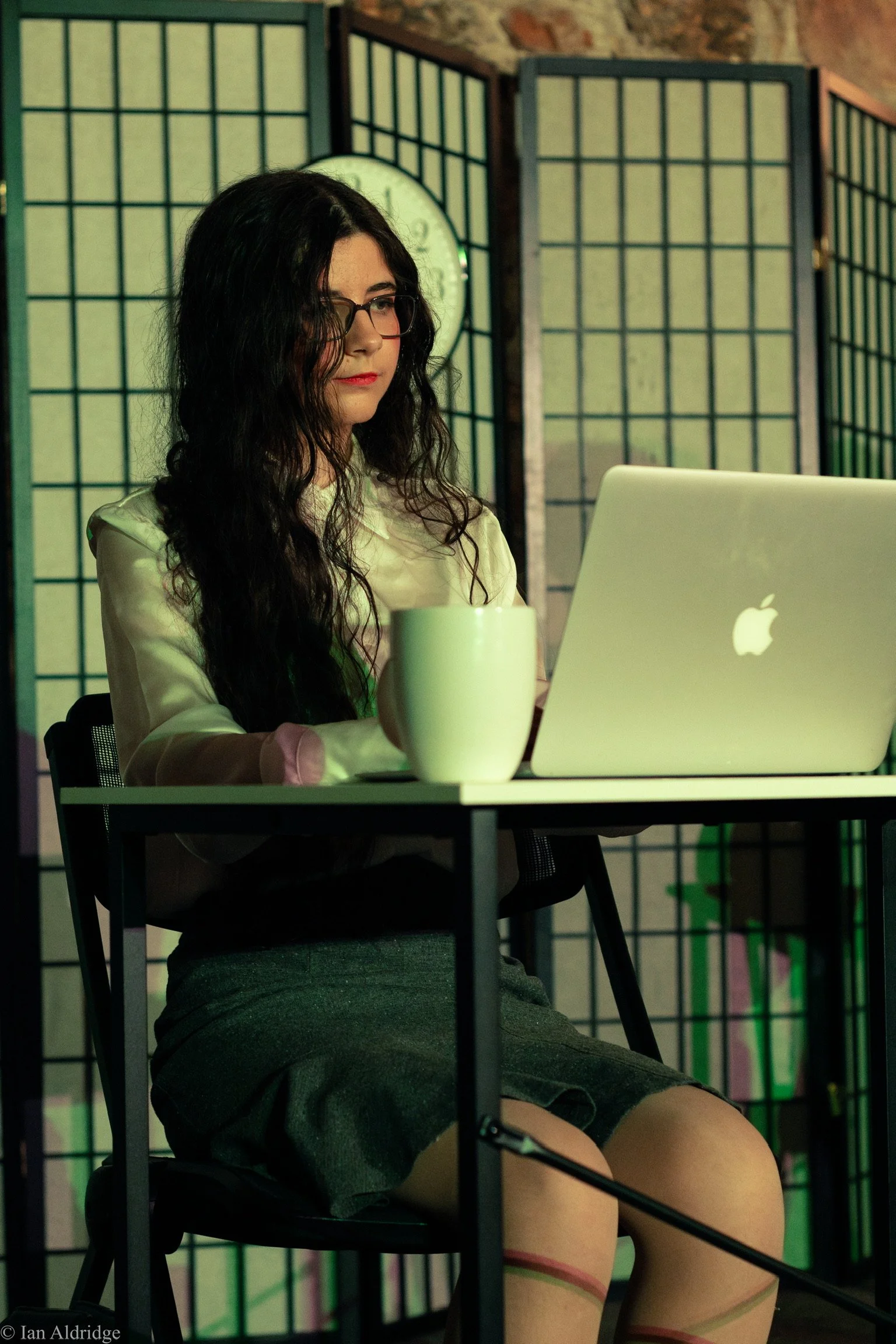 A woman with long black curly hair, wearing glasses, a cream blouse, and gray shorts, is sitting at a black table working on a silver Apple MacBook. Behind her are three folding room dividers with wooden frames and beige panels, and a round clock in 