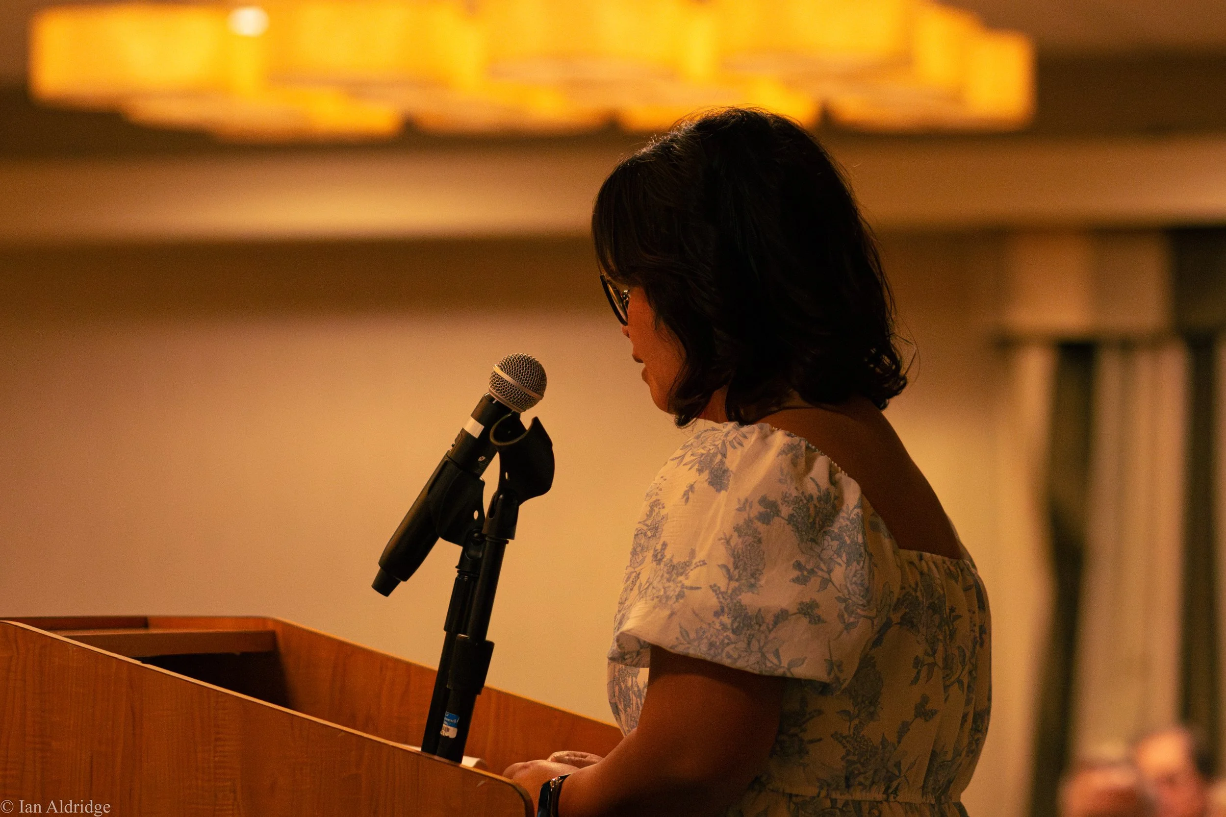 A woman with dark hair and glasses is standing at a podium and speaking into a microphone in a dimly lit room.