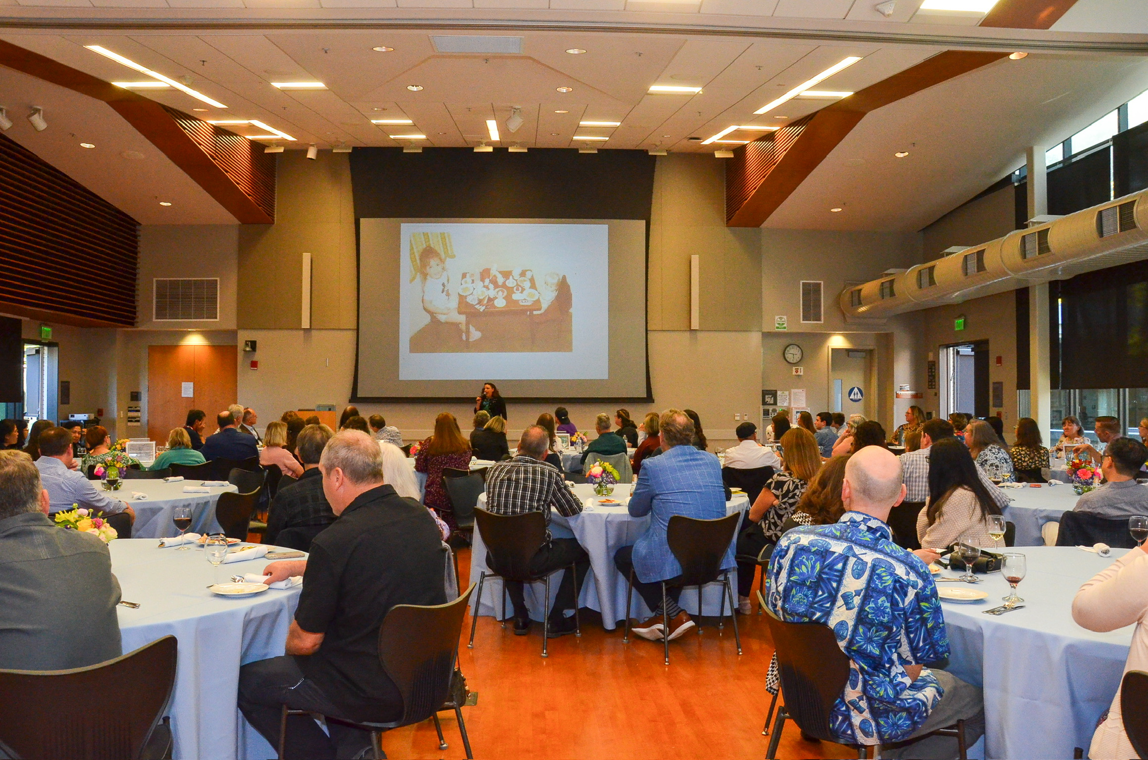 An indoor conference or seminar with many attendees seated at round tables, facing a stage with a speaker and a large screen displaying an image of children playing a board game or eating at a table. The room has a high ceiling, wood accents, and lar