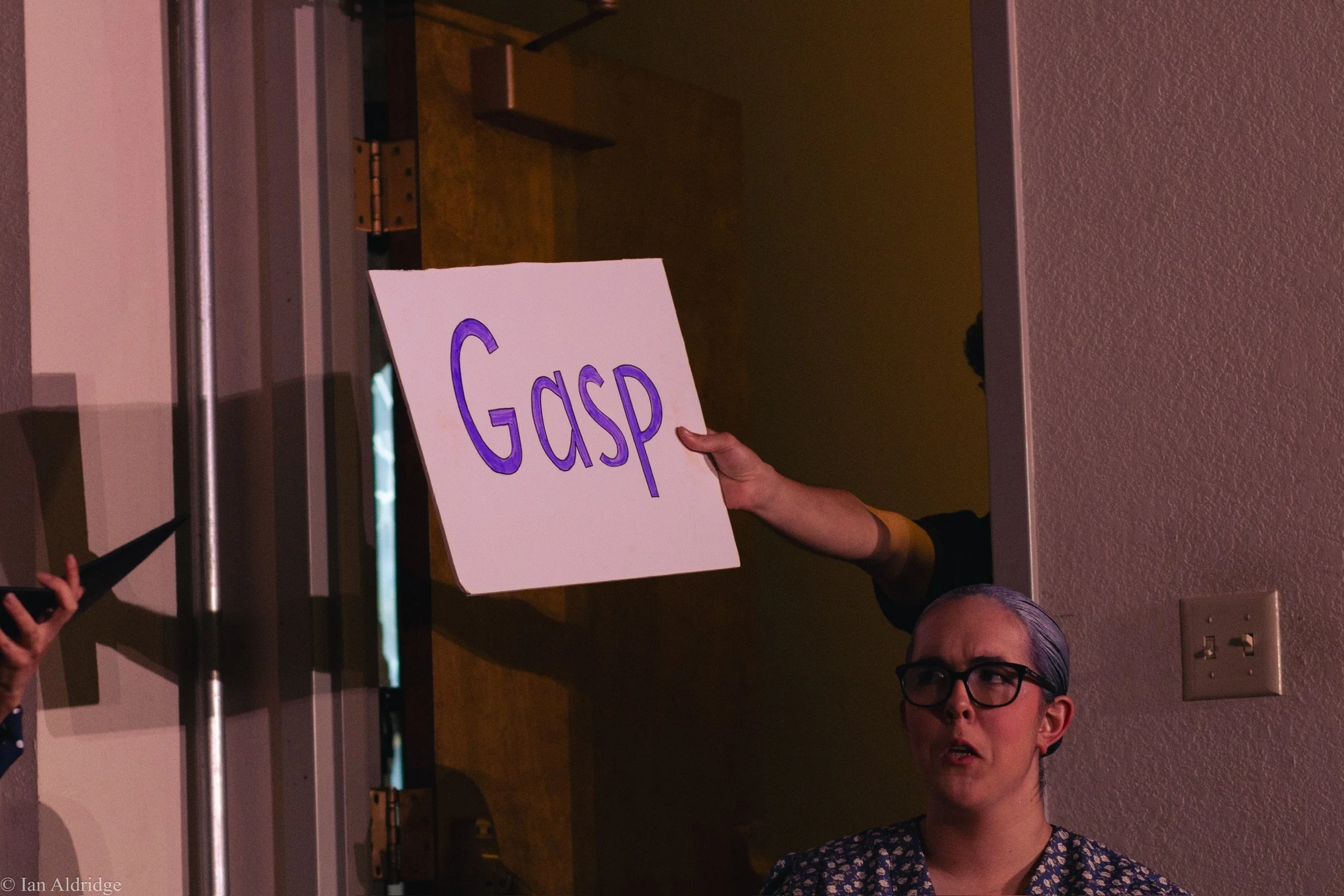 A person holds a white sign with the word 'GASP' written in large purple letters during a gathering or protest at an indoor location.
