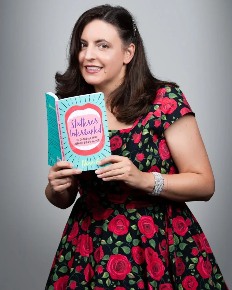 A woman with dark brown hair wearing a black dress with red roses, holding a book titled 'Stutter Interrupted' by Nina G, smiling at the camera against a plain gray background.