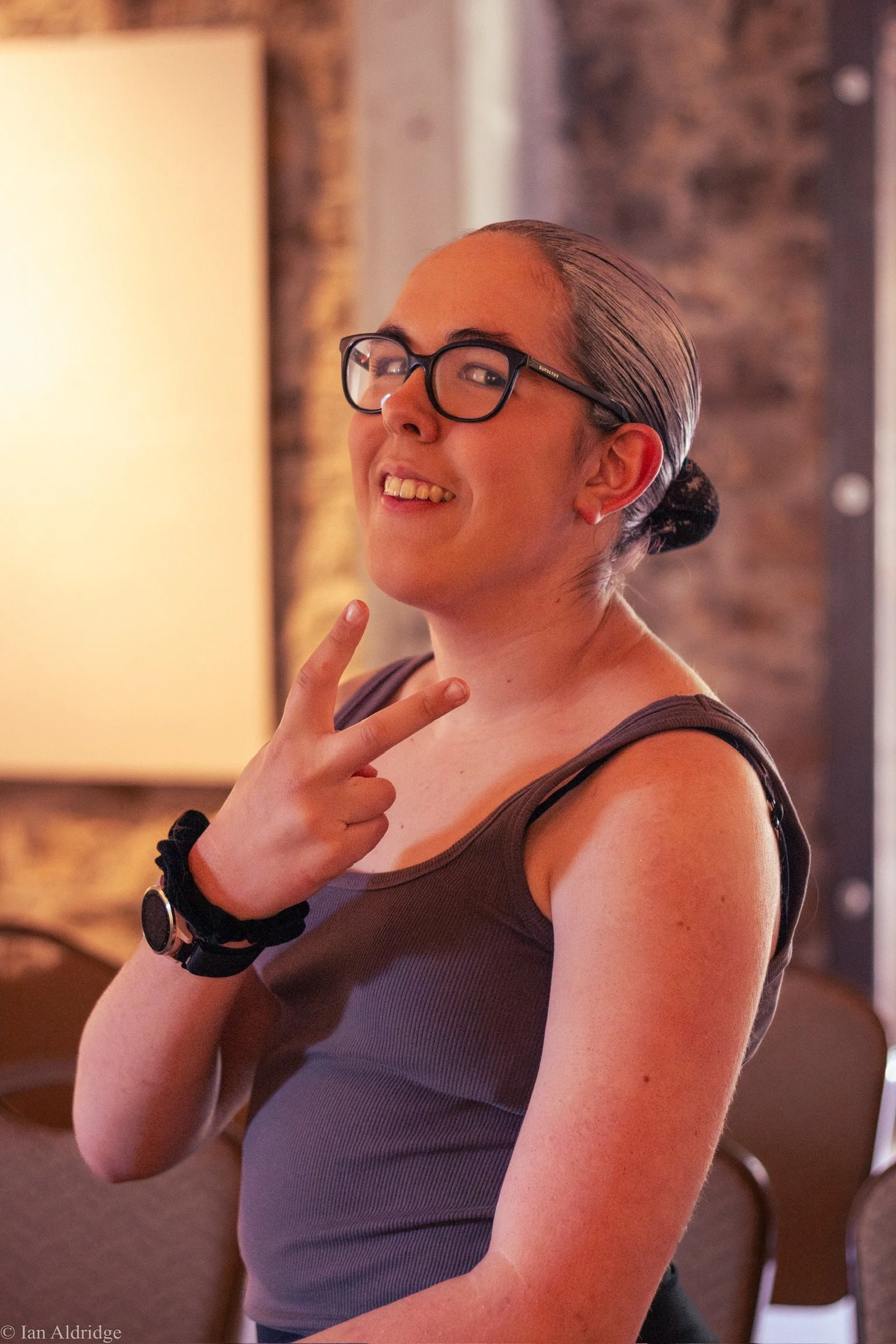 A woman with glasses smiling and making a peace sign gesture with her hand. She has her hair slicked back and is wearing a dark tank top. The background appears to be an indoor space with a brick wall and warm lighting.