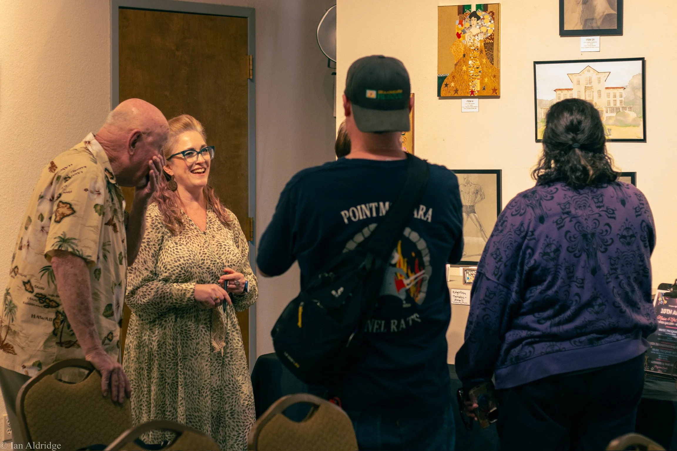 People gathered at an art gallery, viewing framed artwork on the wall. The group includes an elderly man and woman, a woman with red hair and glasses, and a man with a black cap and backpack. The scene appears to be a social event.