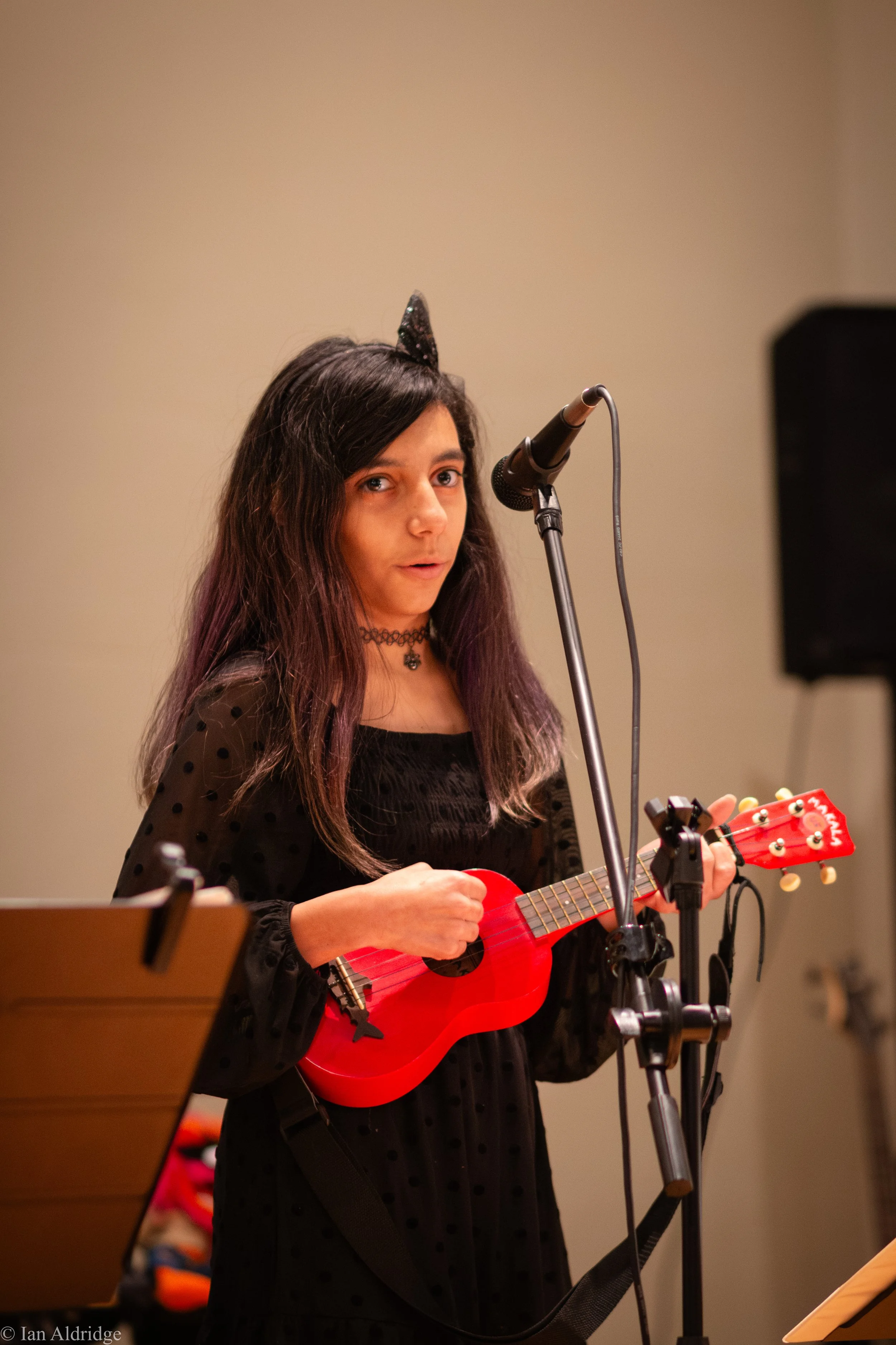 Young woman with long dark hair and a black choker wearing a black dress with polka dots, playing a small red ukulele in front of a microphone on a stand.