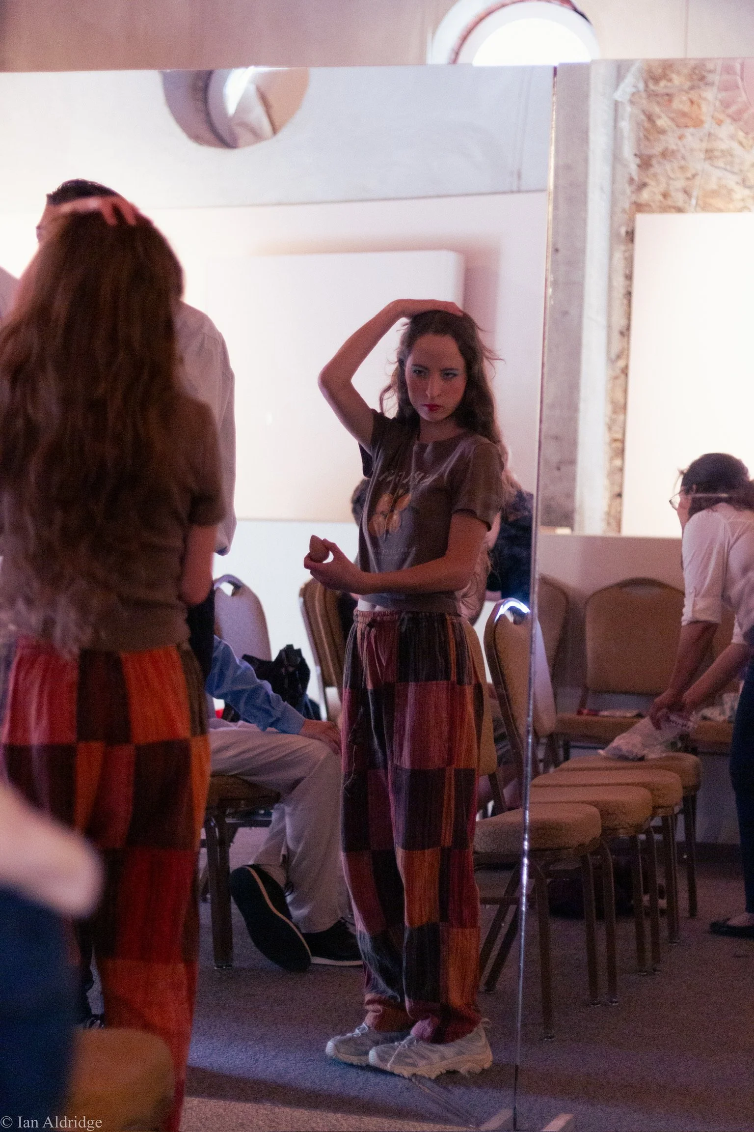 A young woman with long brown hair and red lipstick stands in front of a mirror, touching her hair with her left hand. She is wearing a tan T-shirt and colorful striped pants, and holding a piece of food in her right hand. There are chairs and people