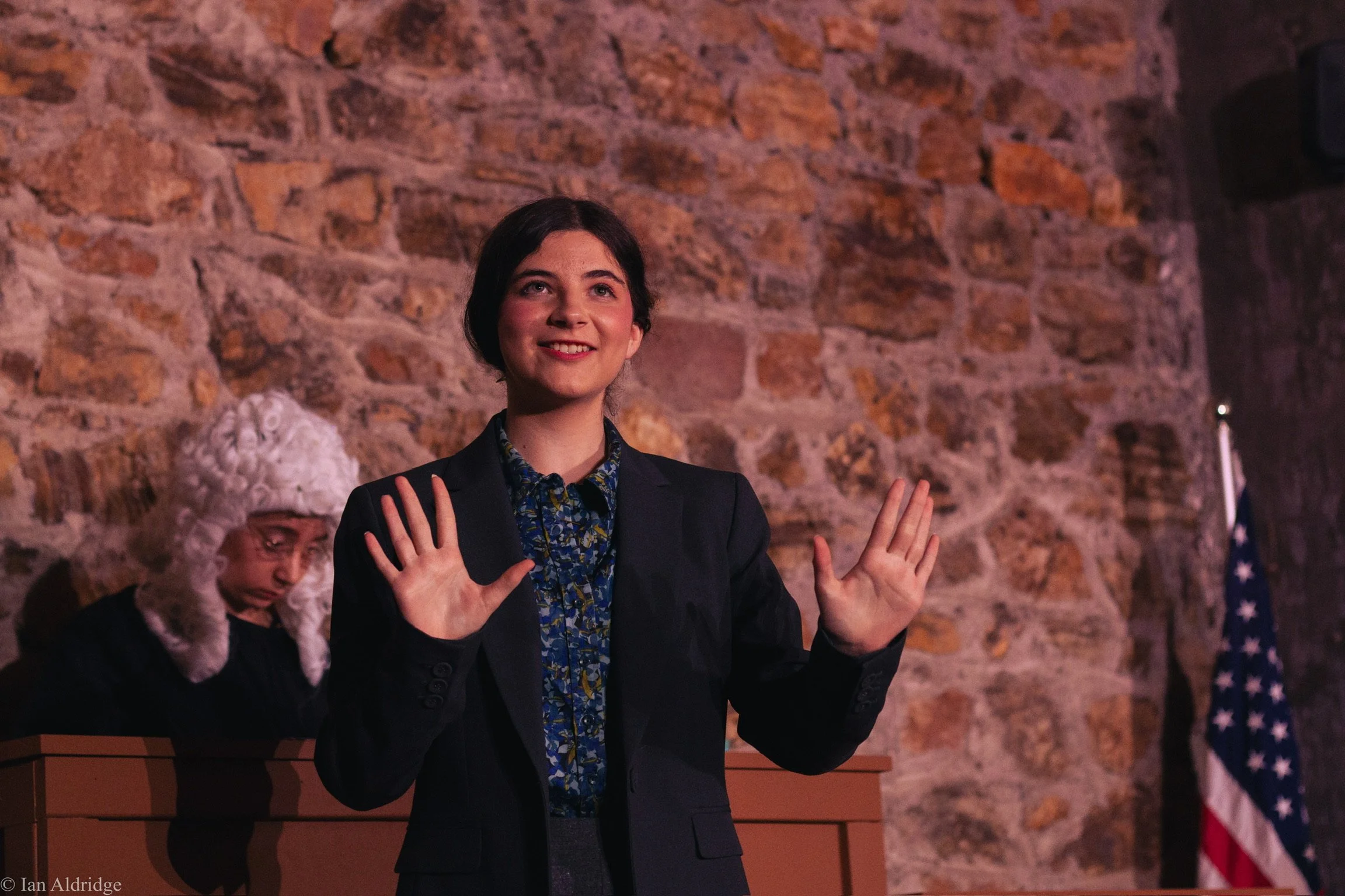A woman with short dark hair smiling and holding her hands up, wearing a dark blazer and a patterned blouse, standing in front of a brick wall. Partially visible to the left is an older woman with white hair and glasses, wearing a black outfit, and b