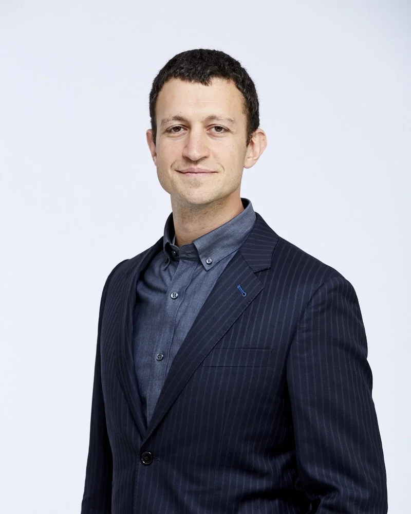 Portrait of a man in a dark pinstripe suit and blue shirt, smiling slightly, against a light background.